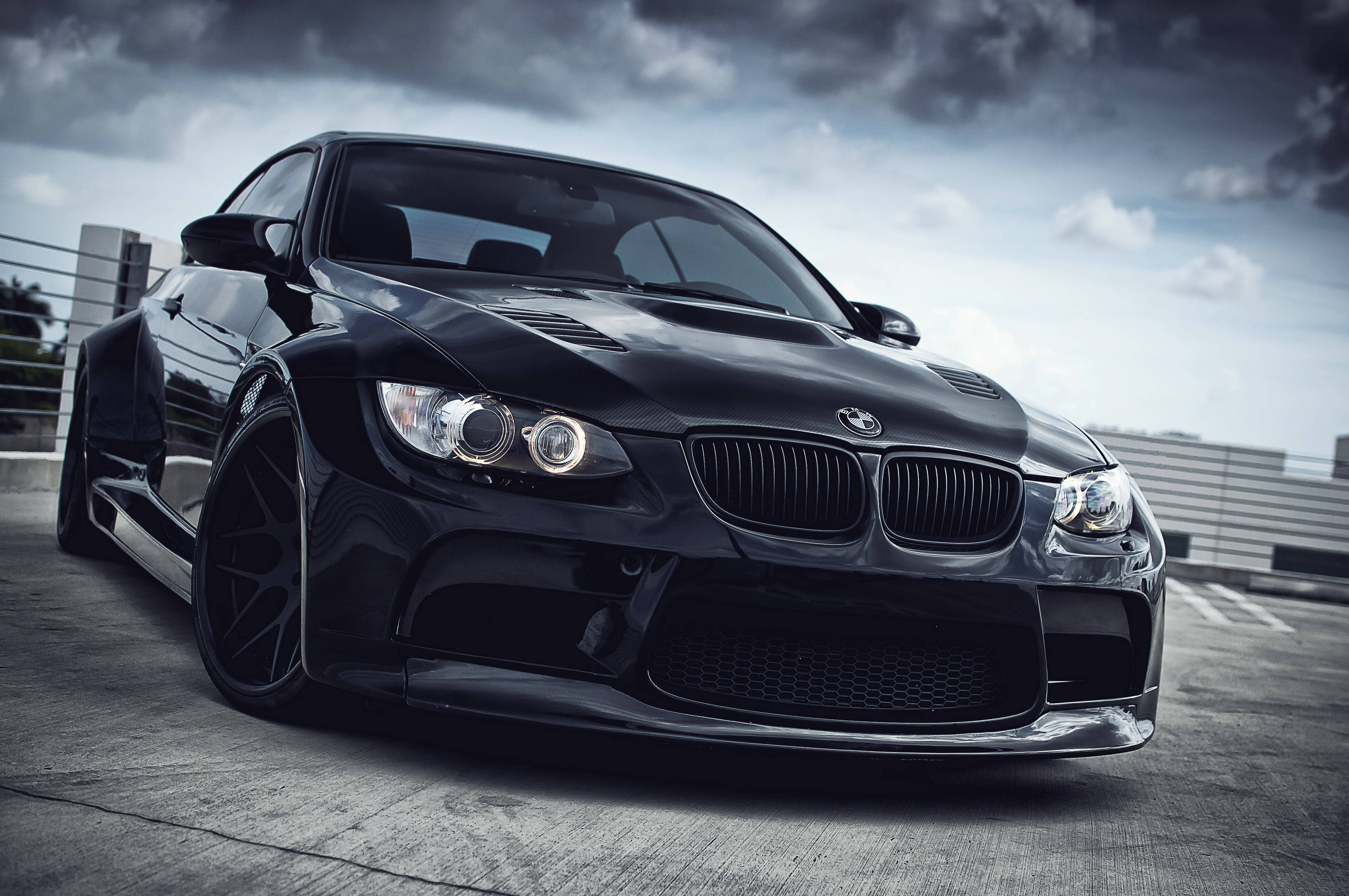 4K BMW 壁紙 桌布 壁紙 — 一架時尚的黑色BMW跑車，配備定制寬體套件和發光的車頭燈，停在混凝土表面上，背景是戲劇性的多雲天空。