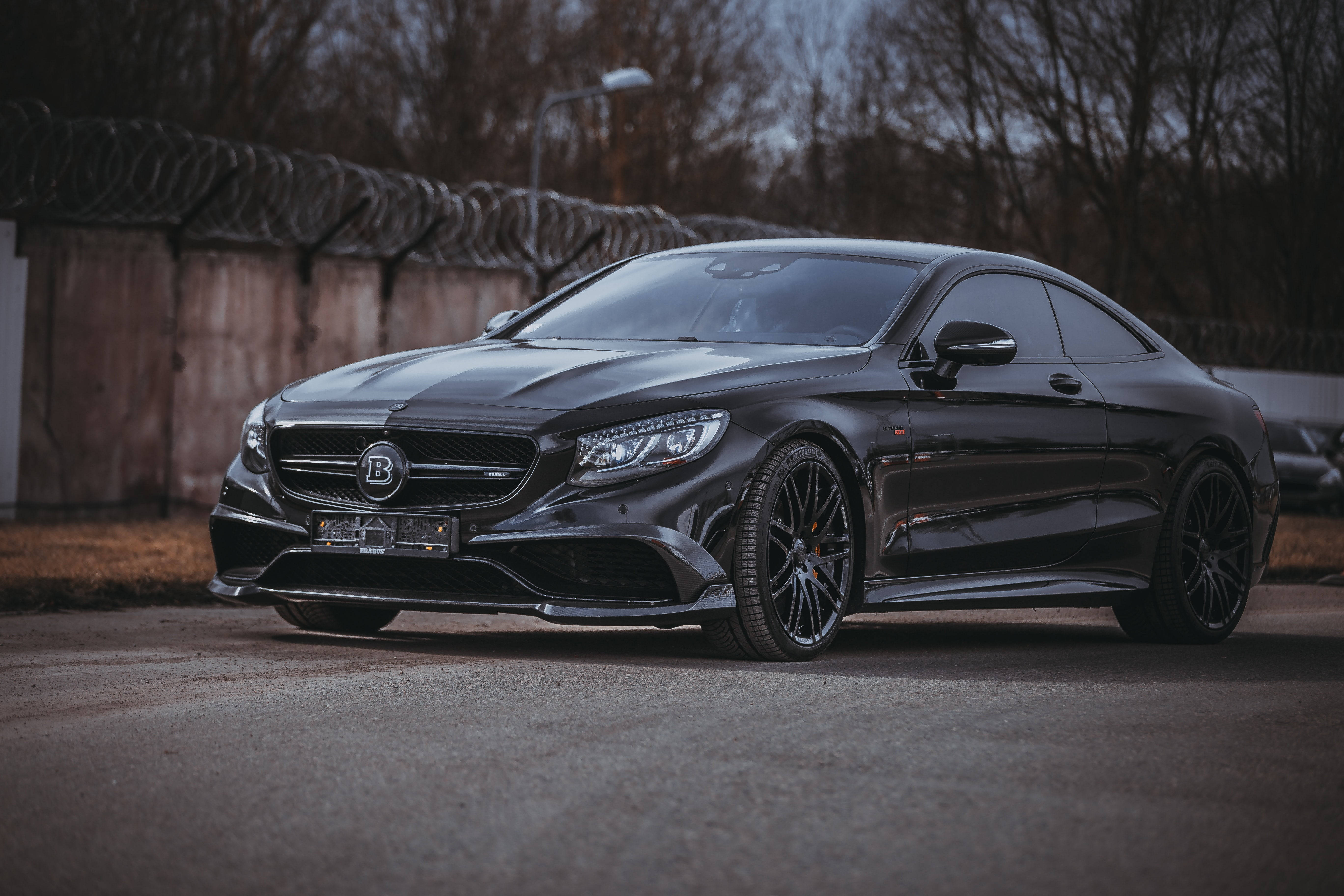 高清 桌布 壁紙 — 一架時尚的黑色Brabus Mercedes-AMG S 65 Coupe停泊在柏油路上，靠近帶刺鐵絲網圍欄，天空多雲。