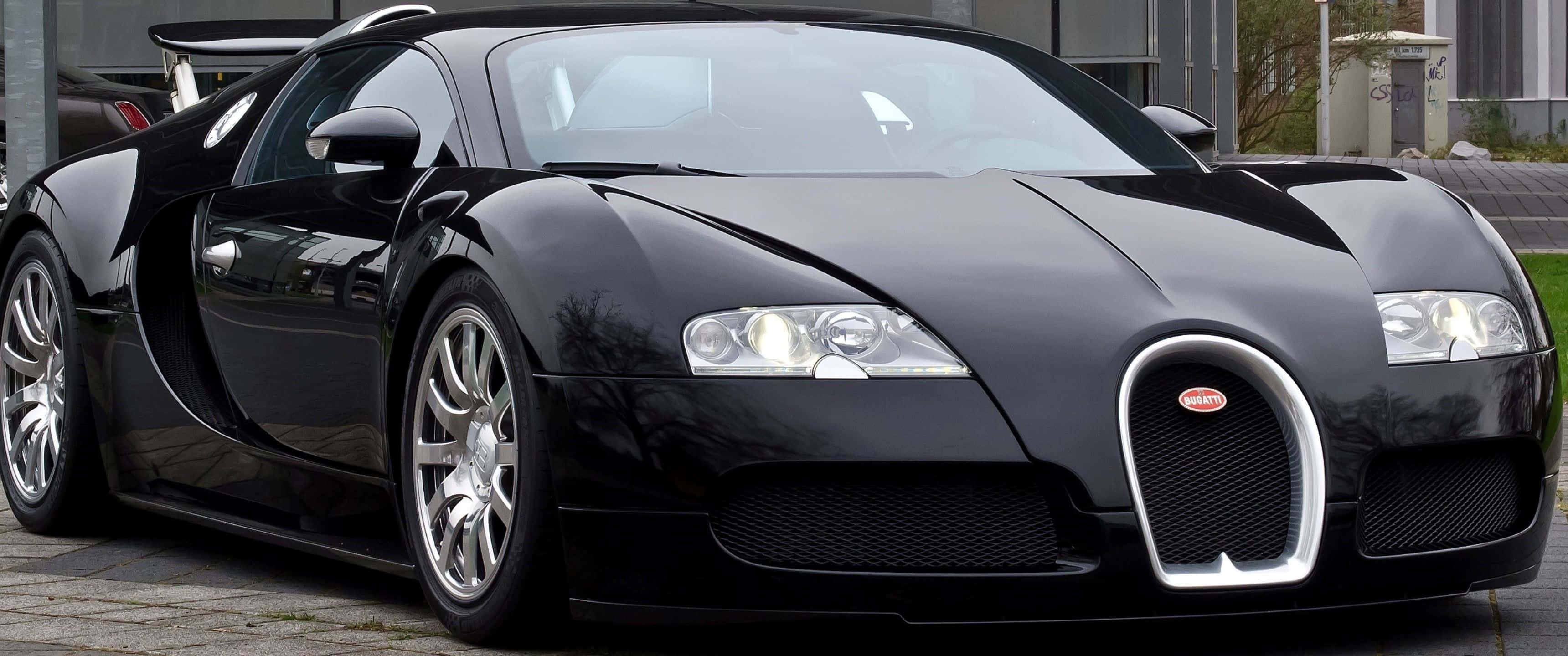 3440x1440p Bugatti 桌布 壁紙 — 一架時尚的黑色Bugatti Veyron停泊在鋪砌的車道上，背景是現代建築。