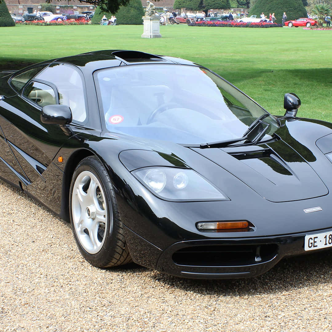 麥拉倫 桌布 壁紙 — 一架時尚的黑色McLaren F1超級跑車在碎石路上閃閃發光，旁邊是修剪整齊的綠色草坪，背景中有人和其他汽車。