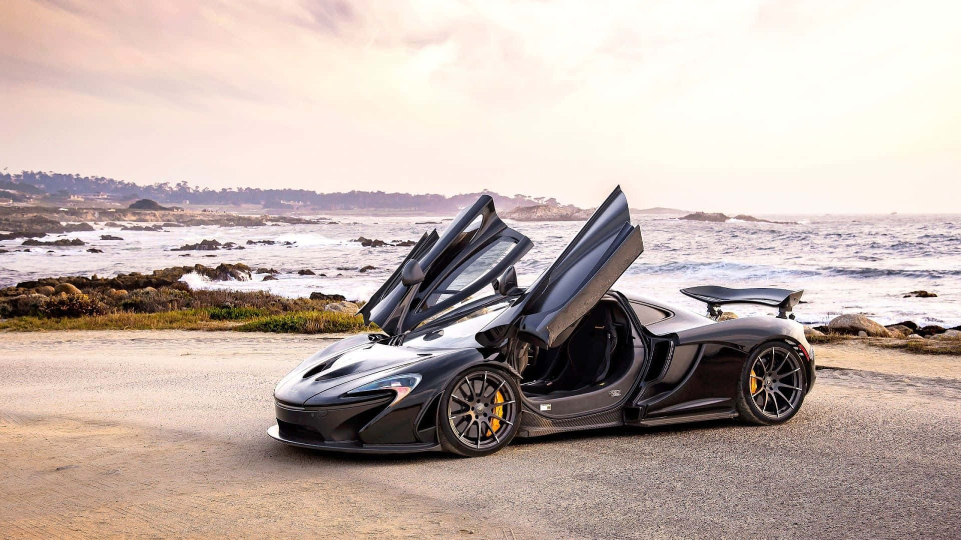 麥拉倫 桌布 壁紙 — 一架時尚的黑色McLaren跑車，鷗翼門打開，停在日落時分的海岸公路上，旁邊是拍岸的波浪。
