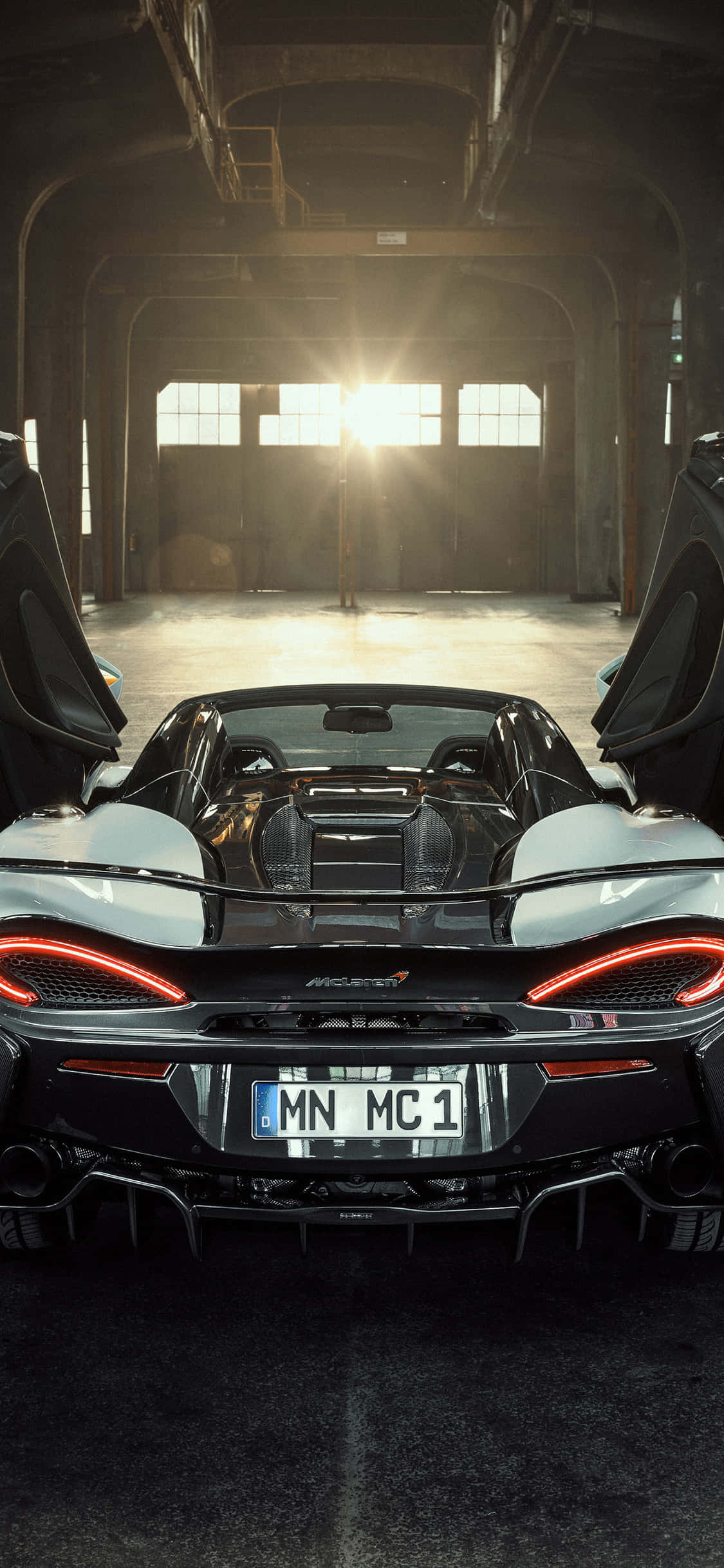 iPhone Xs Mclaren 720s 桌布 壁紙 — 一架時尚的黑色McLaren跑車，帶有發光的紅色尾燈，停放在陽光普照的工業倉庫中，鷗翼車門敞開。