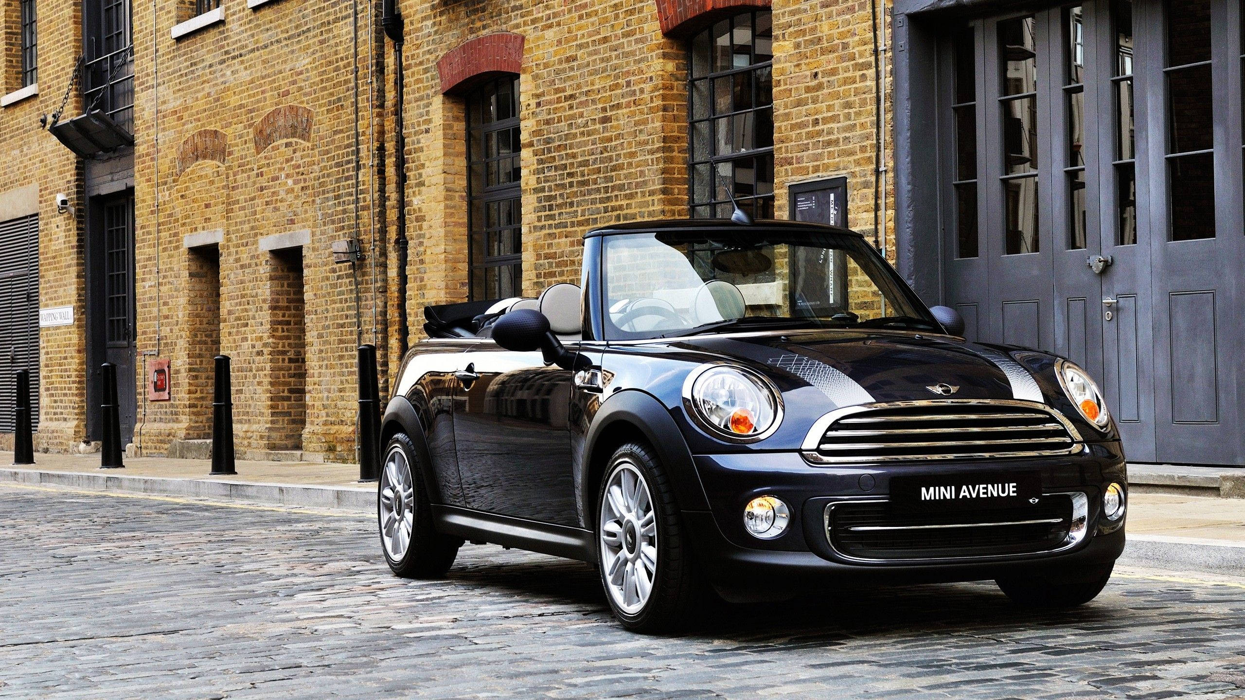 Mini Cooper 桌布 壁紙 — 一架時尚的黑色MINI敞篷車停泊在鵝卵石街道上，旁邊是一座有大型窗戶的風化磚砌建築。