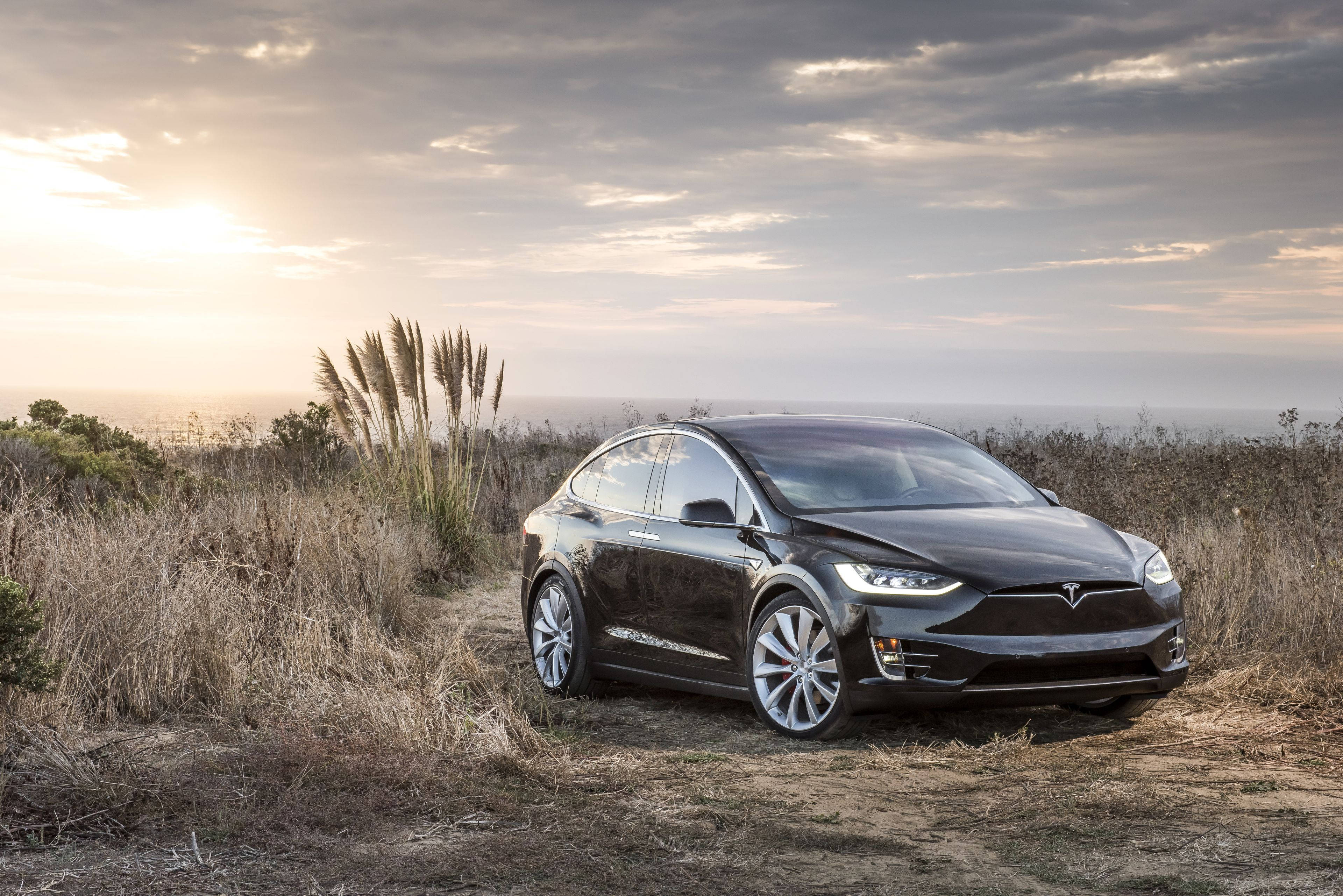特斯拉 桌布 壁紙 — 一架時尚的黑色Tesla Model X停泊在泥濘小徑上，俯瞰日落時分的海洋，金色光芒照亮整個場景。