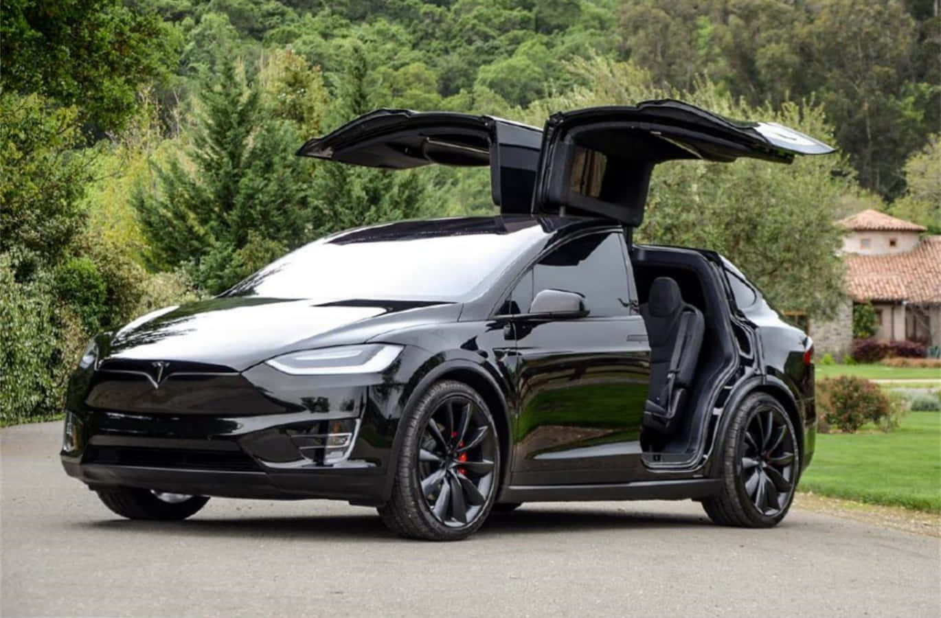 特斯拉 桌布 壁紙 — 一架時尚的黑色Tesla Model X，鷹翼門打開，停在鋪砌的道路上，周圍是茂密的綠樹和石屋。