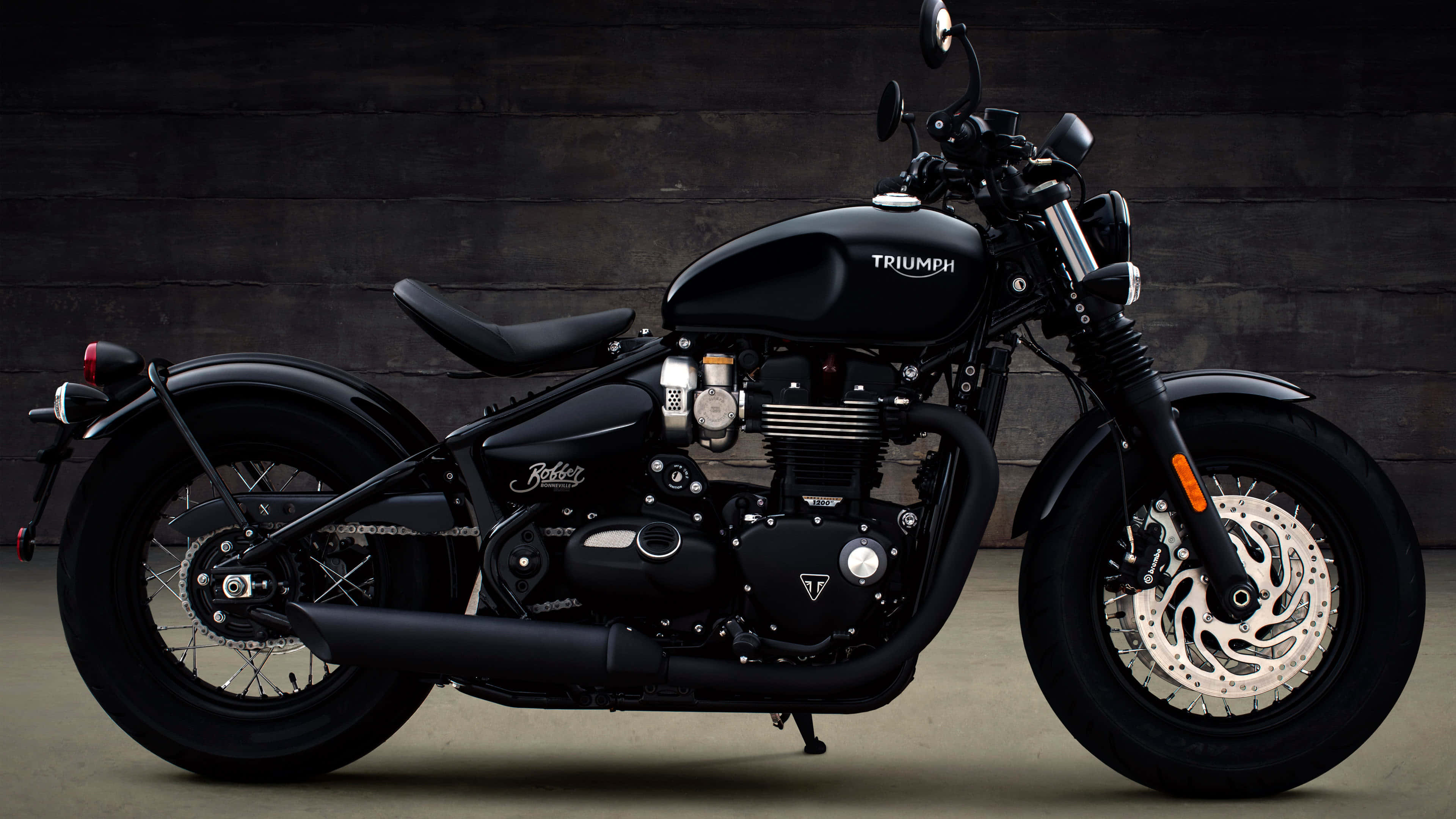 4K Bikes 桌布 壁紙 — 一架時尚的黑色Triumph機車靠著深色木牆，展示其鍍鉻裝飾和堅固設計。