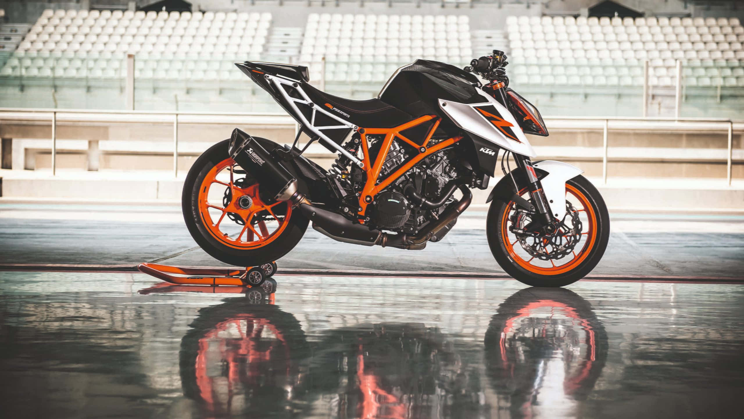 Ktm Bike 桌布 壁紙 — 一架時尚的黑色和橙色KTM摩托車停在一個空蕩蕩的體育館內，地面濕滑反光，周圍是白色座位。