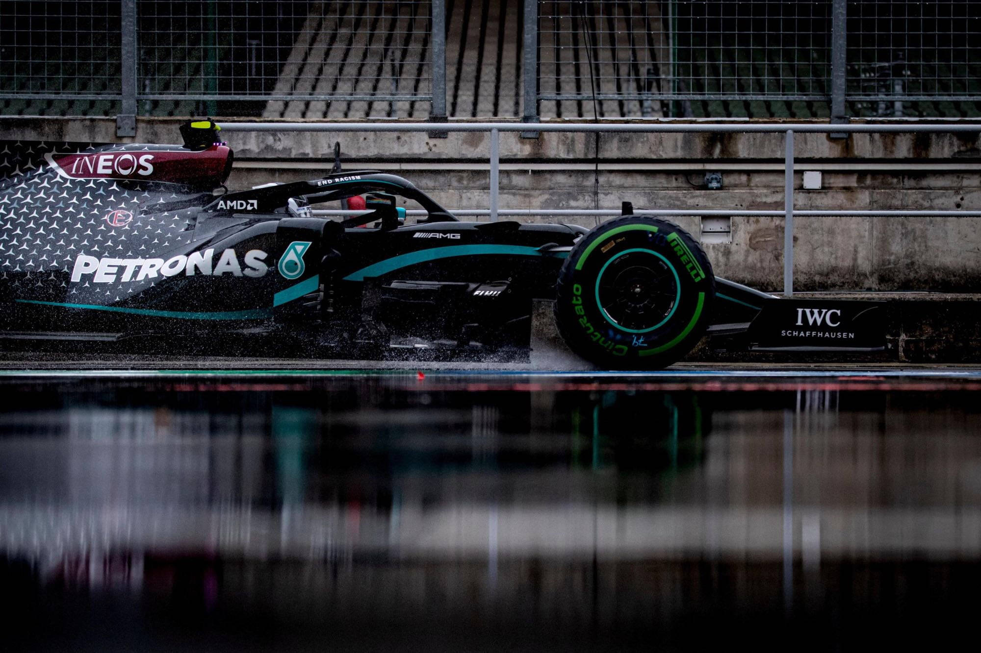 F1 桌布 壁紙 — 一架時尚的黑色和藍綠色一級方程式賽車在潮濕賽道上濺起積水，背景有混凝土護欄和圍欄。