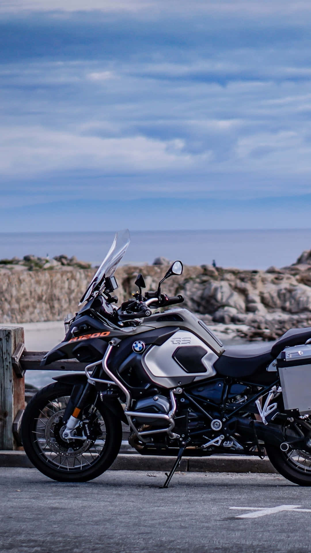 iPhone 桌布 壁紙 — 一架時尚的黑色和銀色BMW R 1250 GS冒險摩托車停泊在沿海道路上，旁邊有岩石懸崖和海洋，天空多雲。