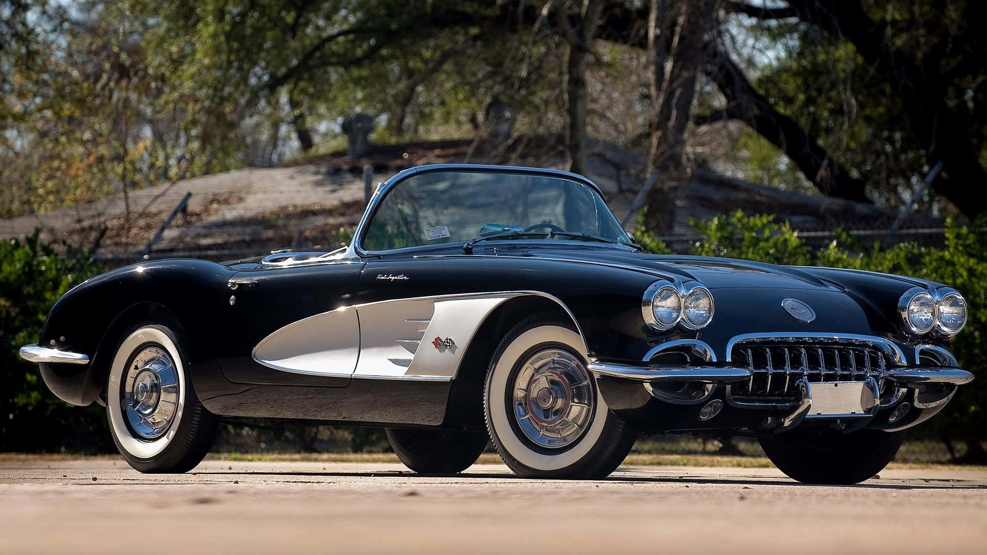 classic 桌布 壁紙 — 一架時尚的黑色和銀色經典Corvette跑車停泊在鋪砌的道路上，在明亮的陽光下，背景有樹木。