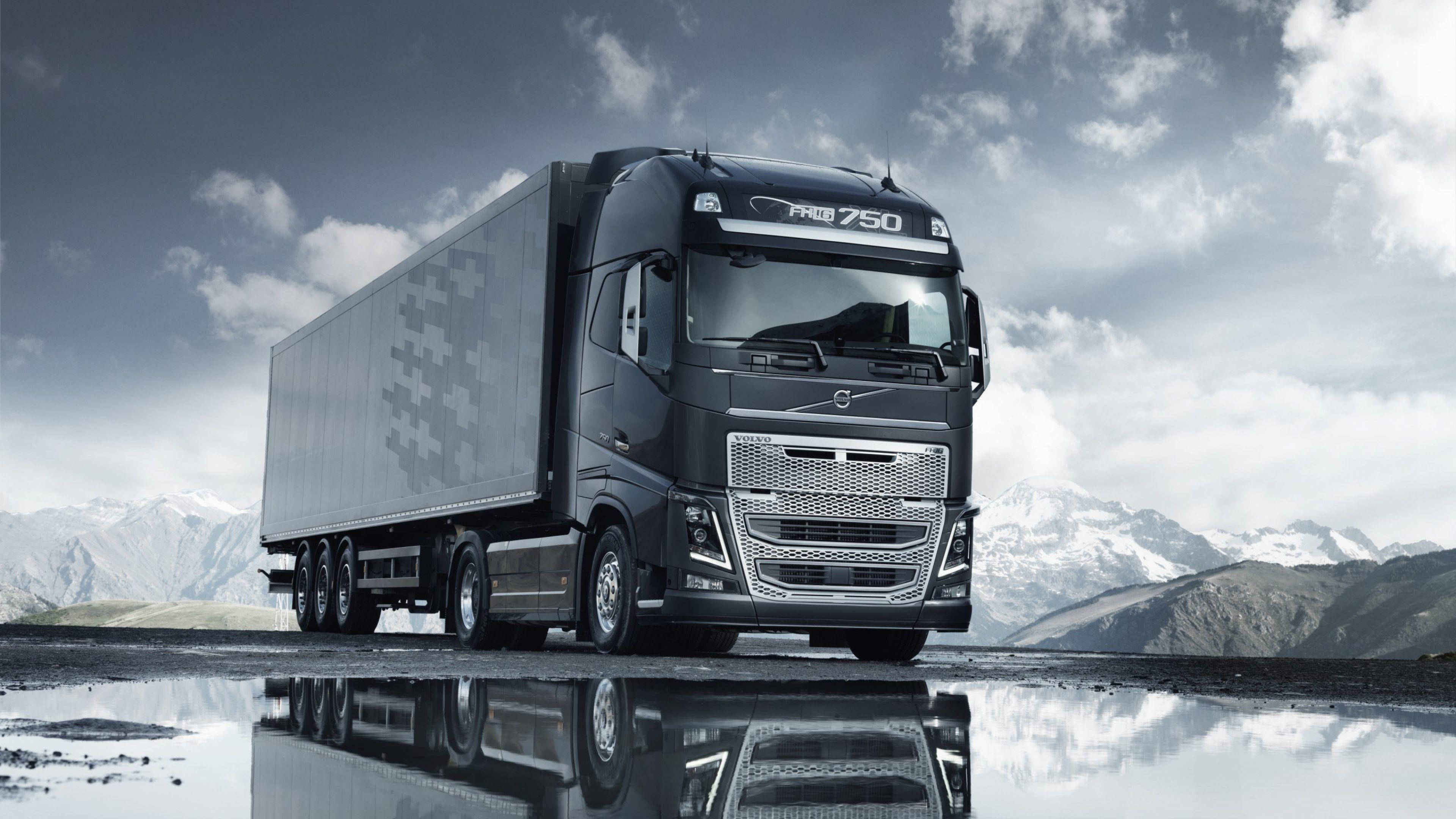 Trucks 桌布 壁紙 — 一架時尚的黑色富豪 FH16 750 卡車，帶有像素化拖車，停在潮濕的道路上，在陰天背景下，映照著積雪覆蓋的山脈。