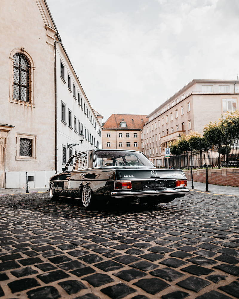 Mercedes 桌布 壁紙 — 一架時尚的黑色復古賓士轎車停泊在歷史悠久的歐洲建築之間的鵝卵石街道上，天空多雲。