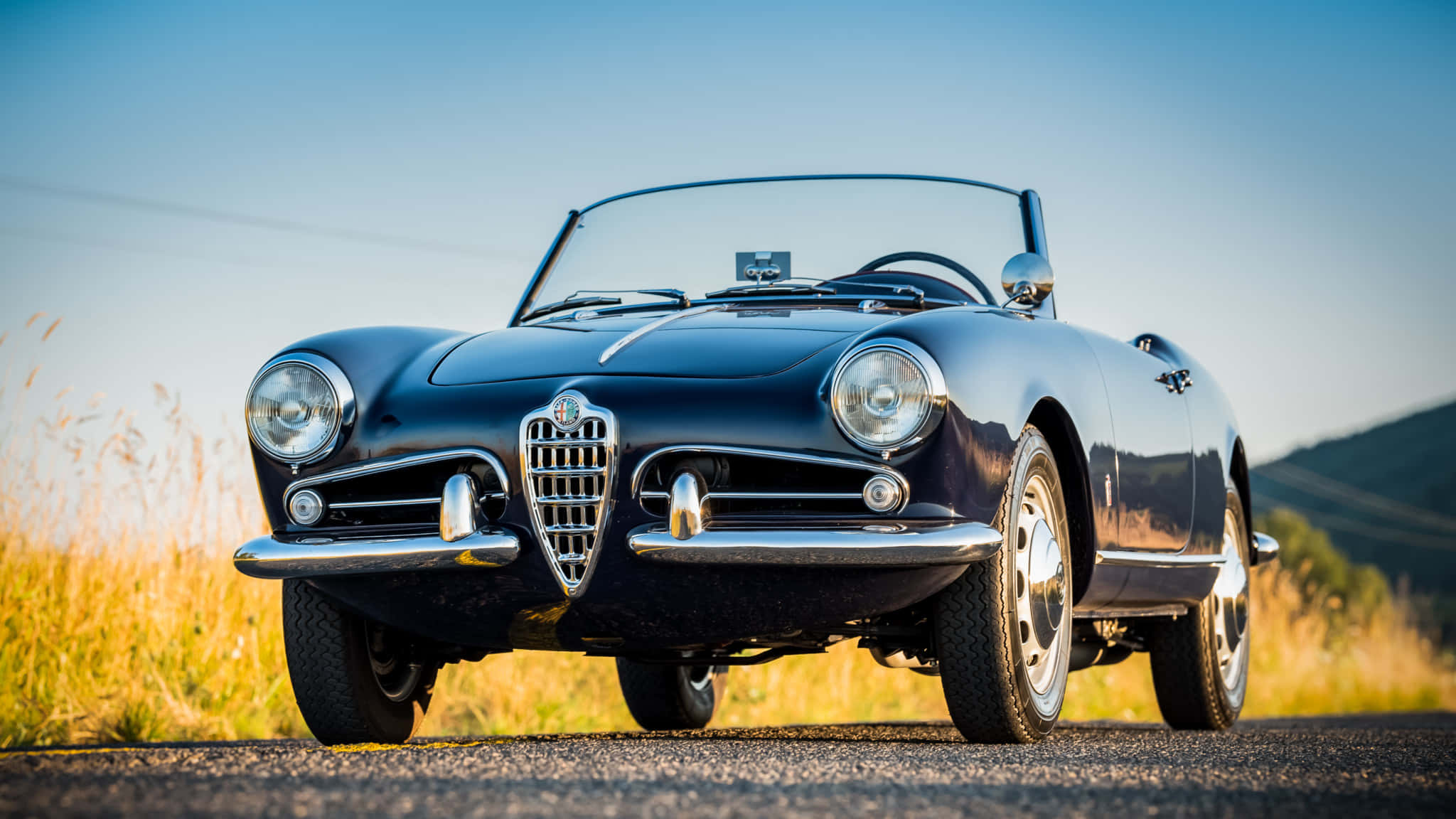 Alfa Romeo Spider 桌布 壁紙 — 一架時尚的黑色愛快羅密歐Spider敞篷車停泊在陽光明媚的鄉村道路上，周圍是金色的草地和遠處的山丘，天空清澈湛藍。