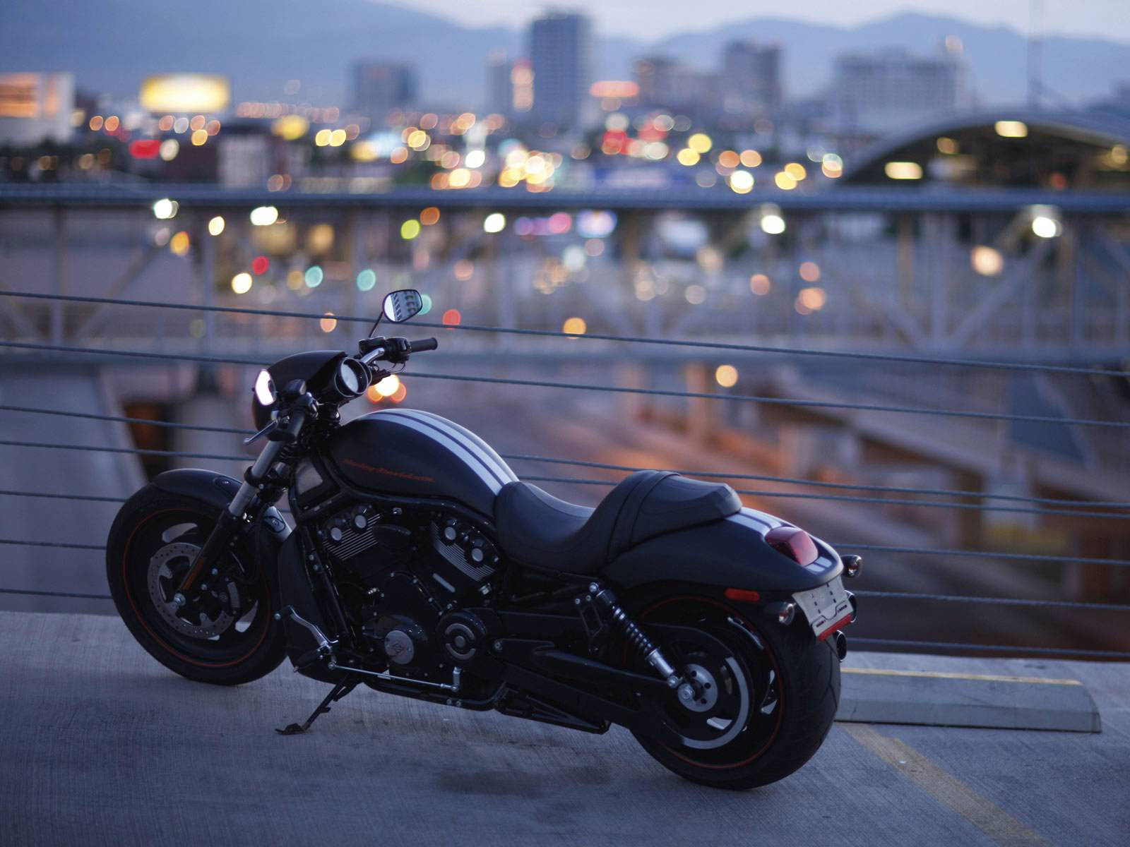 Harley 桌布 壁紙 — 一架時尚的黑色摩托車停放在屋頂上，俯瞰著閃爍著暮色散景燈光的城市天際線。