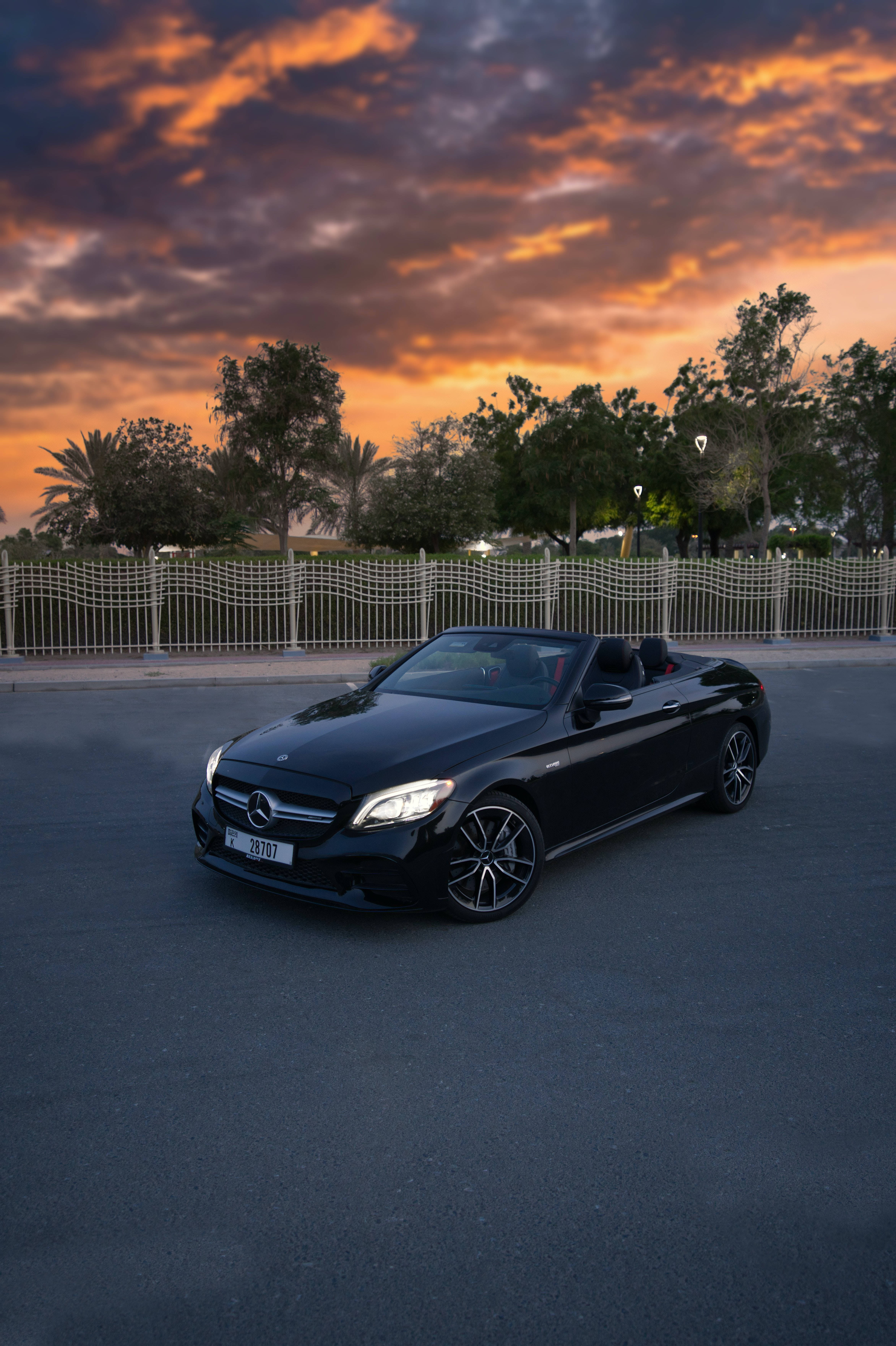 mercedes amg iPhone 桌布 壁紙 — 一架時尚的黑色敞篷跑車停在柏油路上，背景是戲劇性的橙紫色日落天空。