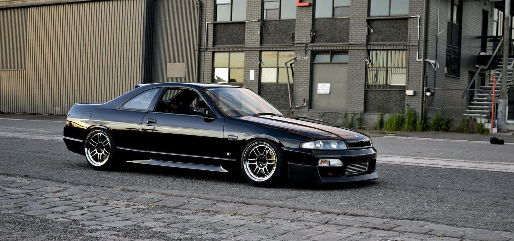 R33 GT-R 桌布 壁紙 — 一架時尚的黑色日產Skyline雙門跑車停泊在工業建築旁的鵝卵石街道上。