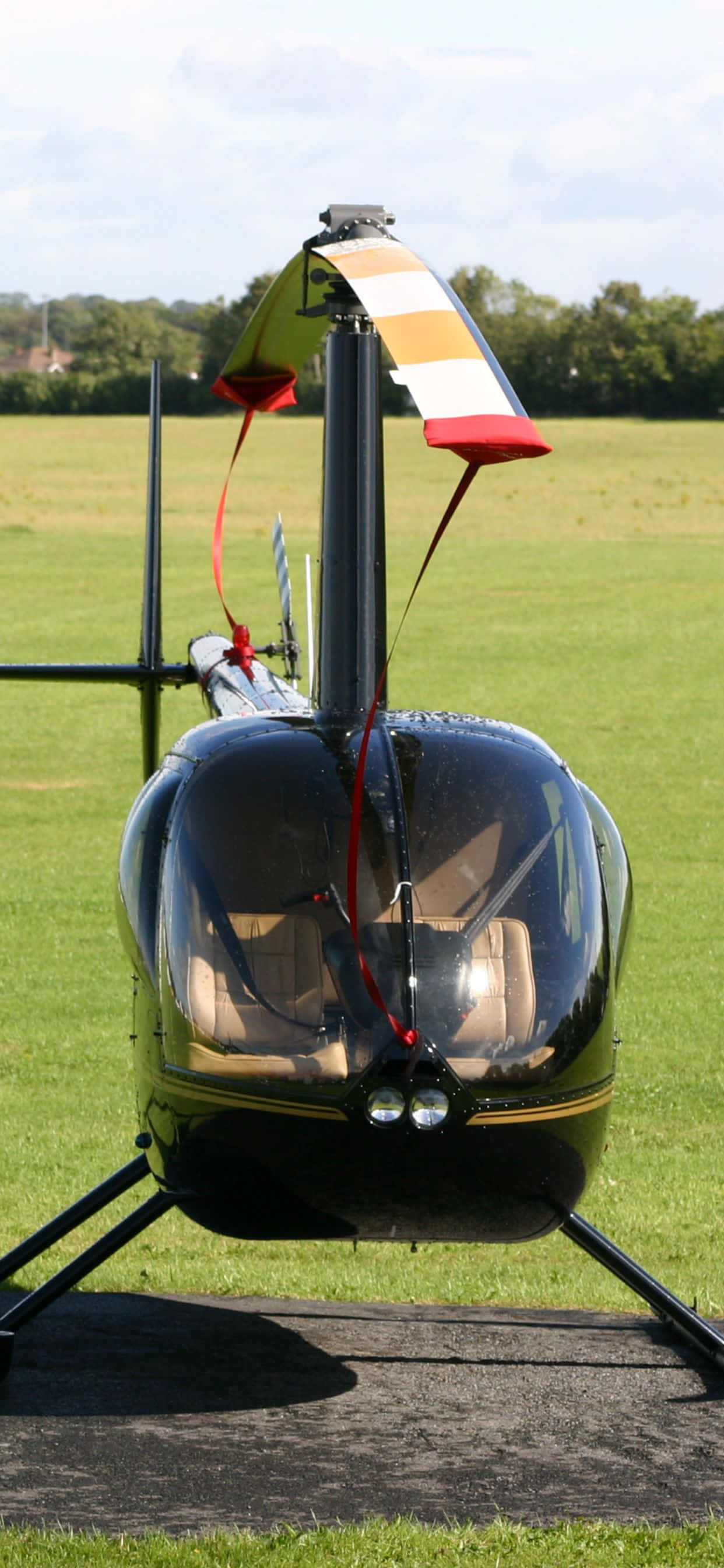 iPhone Xs Max Helicopters 桌布 壁紙 — 一架時尚的黑色直升機帶有彩色尾翼條紋，停在廣闊綠色田野中的鋪砌停機坪上，天空明亮。
