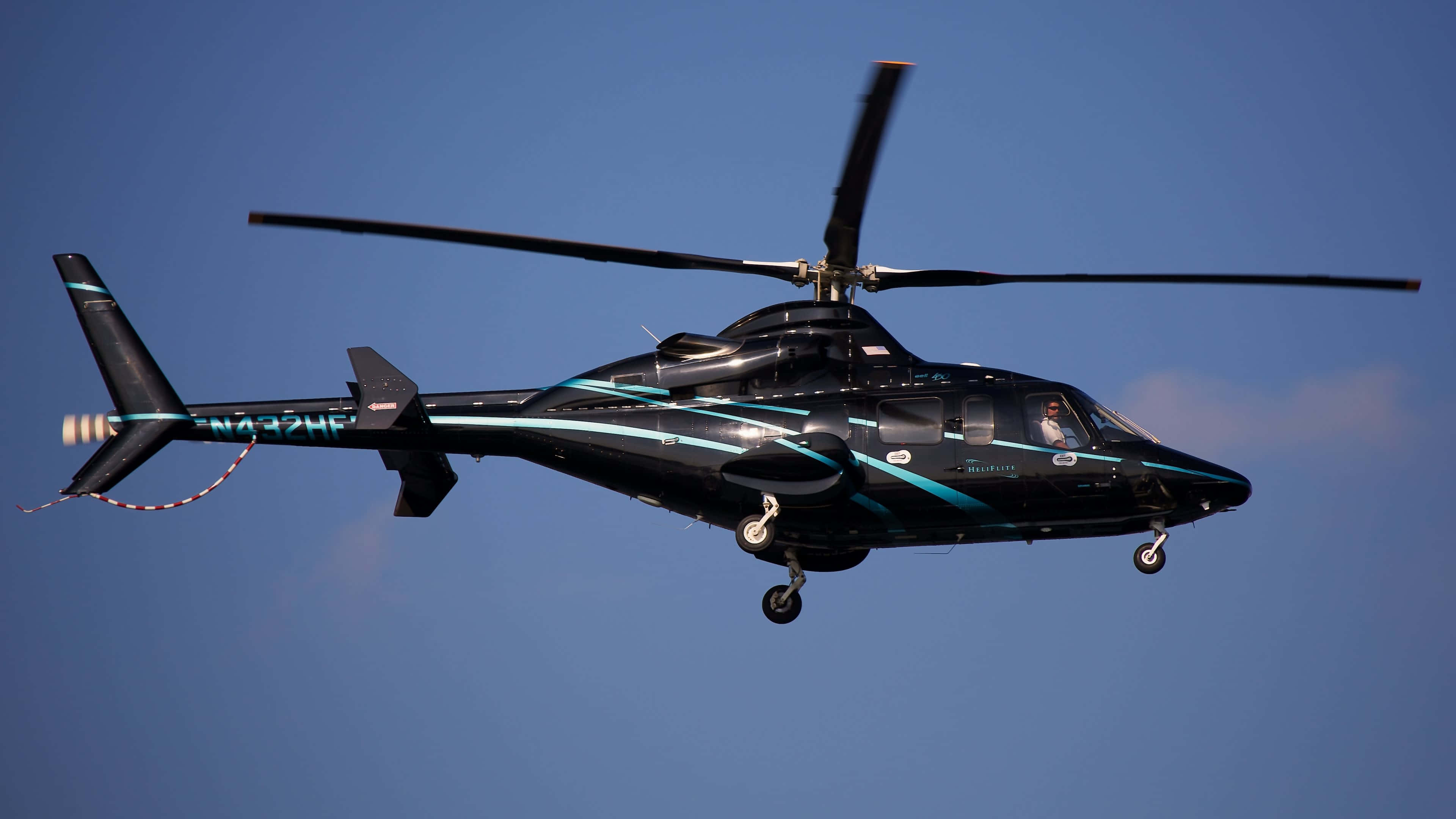4K Helicopters 桌布 壁紙 — 一架時尚的黑色直升機帶有青綠色點綴，放下起落架在晴朗的藍天低飛。