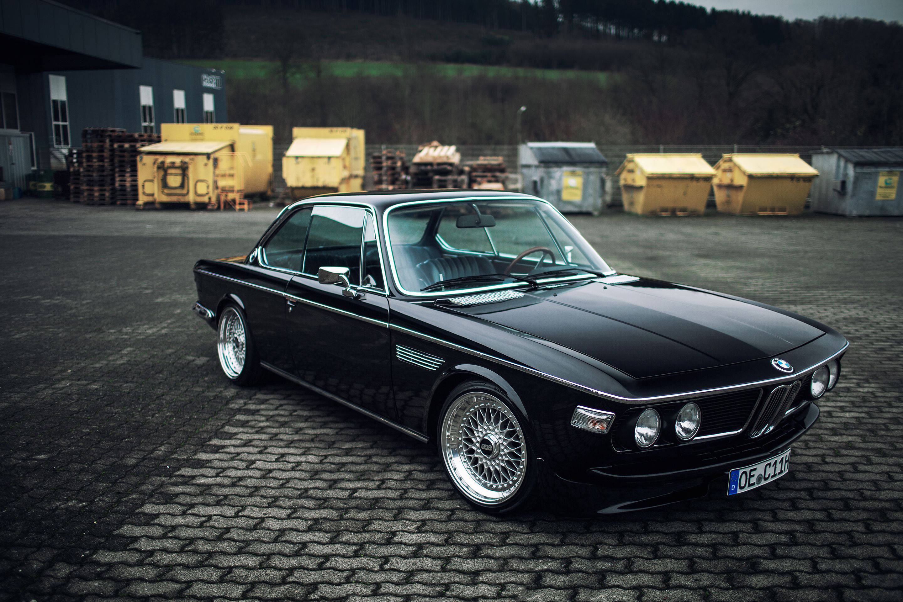 classic bmw 桌布 壁紙 — 一架時尚的黑色經典BMW雙門轎跑車配備鋼絲輪圈，停泊在鵝卵石地上，旁邊是黃色垃圾車，天空多雲。