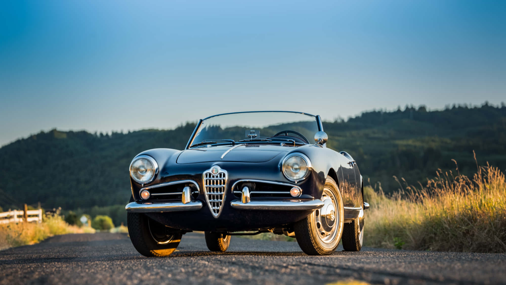 Alfa Romeo Spider 桌布 壁紙 — 一架時尚的黑色經典愛快羅密歐Spider敞篷車停泊在鄉村道路上，背景是連綿起伏的綠色山丘和晴朗的藍天。