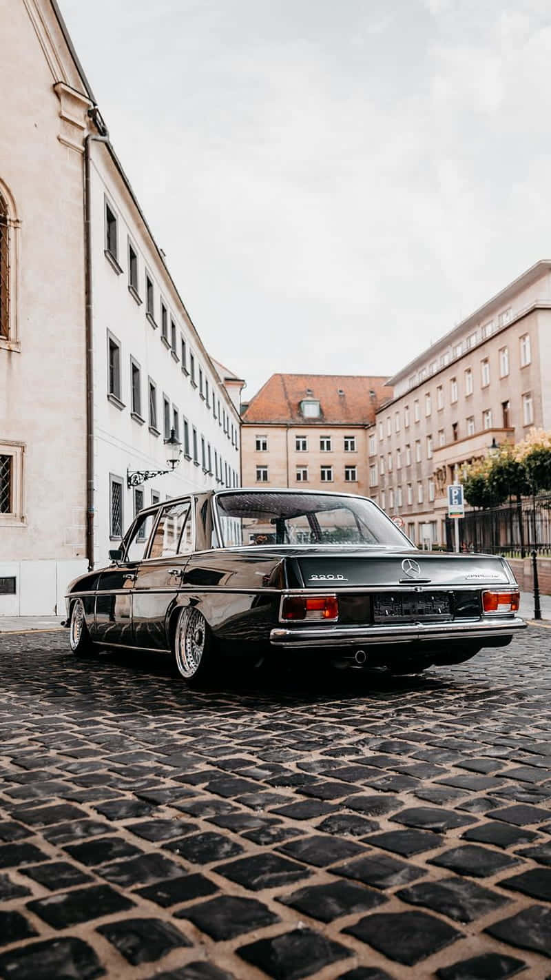 Classic Mercedes 桌布 壁紙 — 一架時尚的黑色經典梅賽德斯-奔馳2800停泊在歷史悠久的歐洲建築之間的鵝卵石街道上，天空多雲。