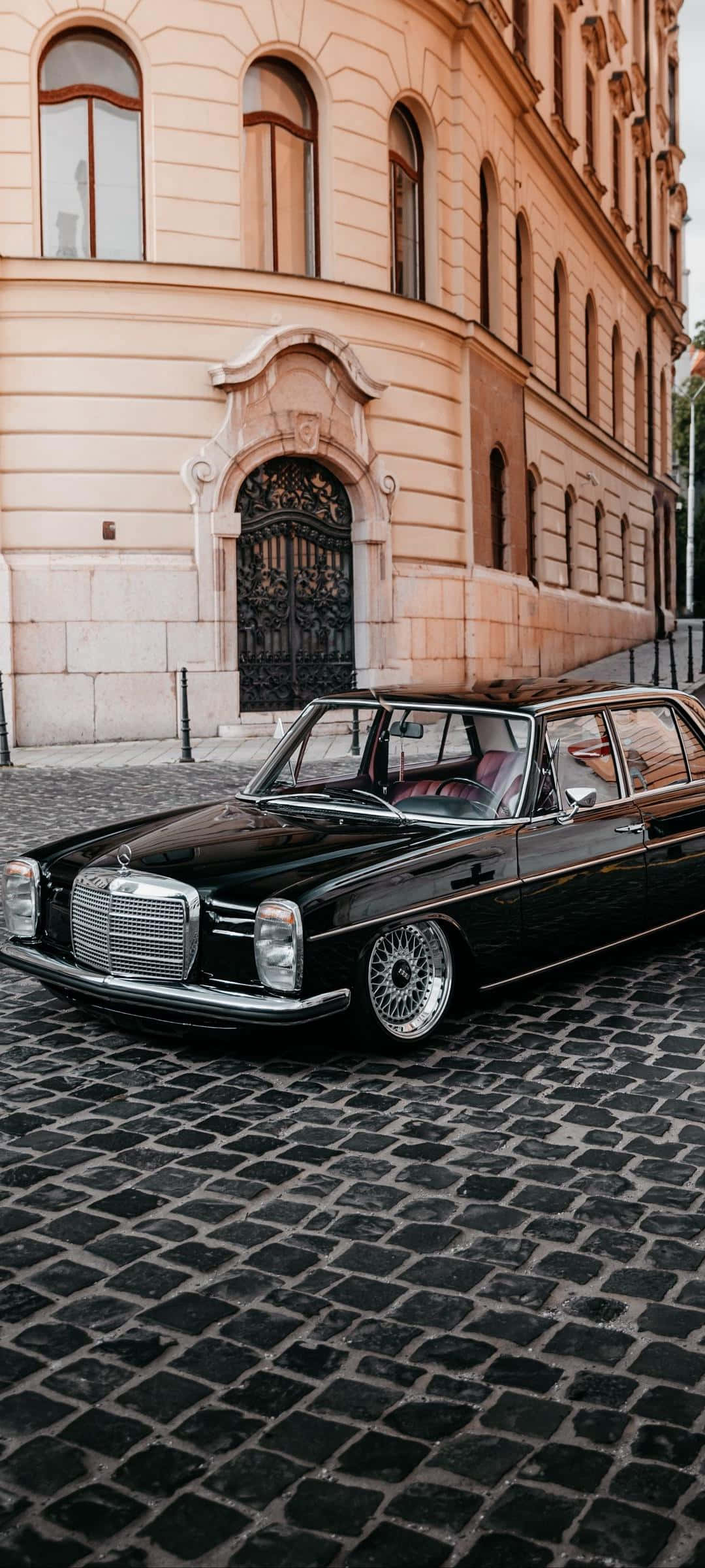 Classic Mercedes 桌布 壁紙 — 一架時尚的黑色經典賓士轎車停泊在鵝卵石街道上，旁邊是一座帶有拱形窗戶的大型桃色建築。