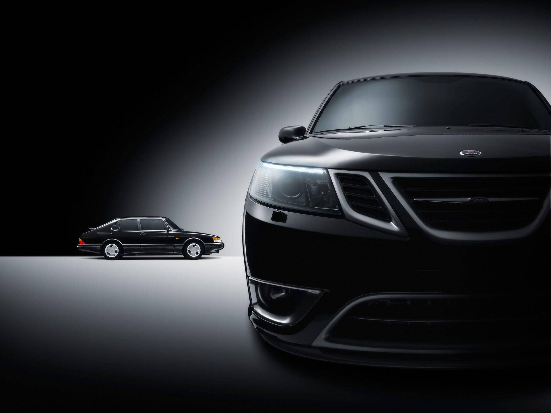 Saab 桌布 壁紙 — 一架時尚的黑色薩博汽車在深色漸變背景上戲劇性地打光，旁邊有一輛較小的復古薩博模型。
