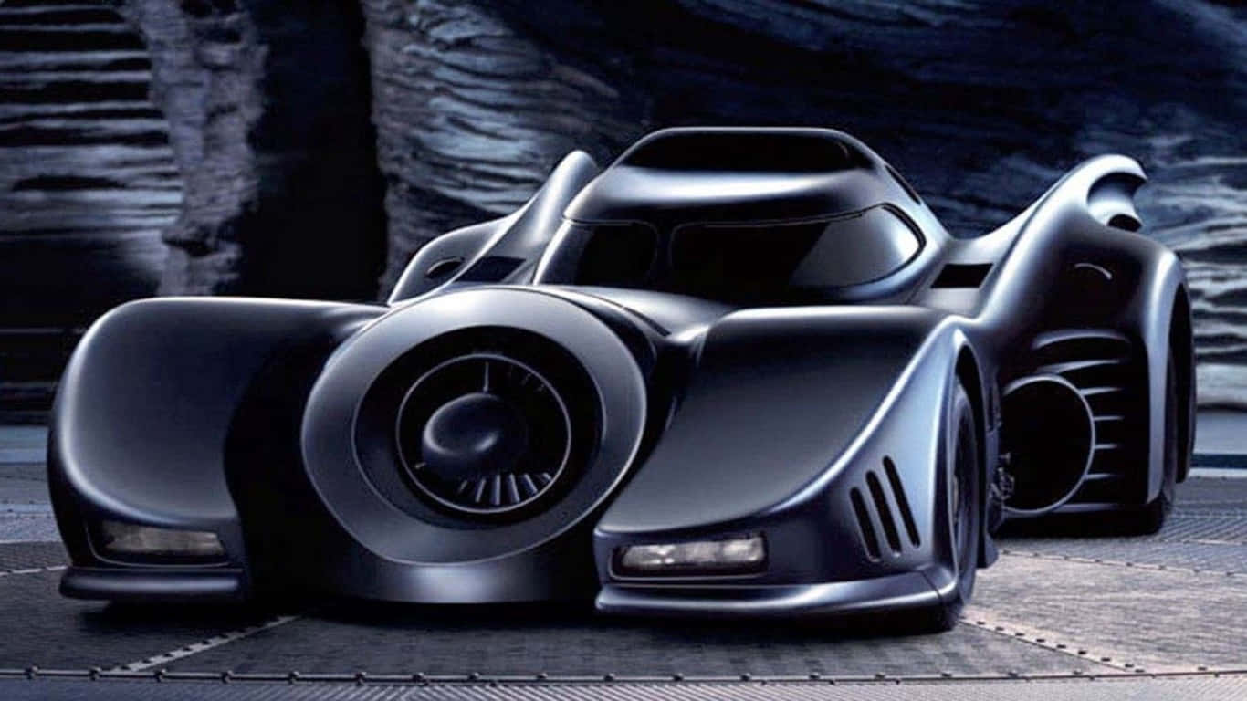 1366x768 Batmobile 桌布 壁紙 — 一架時尚的黑色蝙蝠車，帶有發光的車頭燈，停在一個有紋理石牆的黑暗洞穴般的環境中。