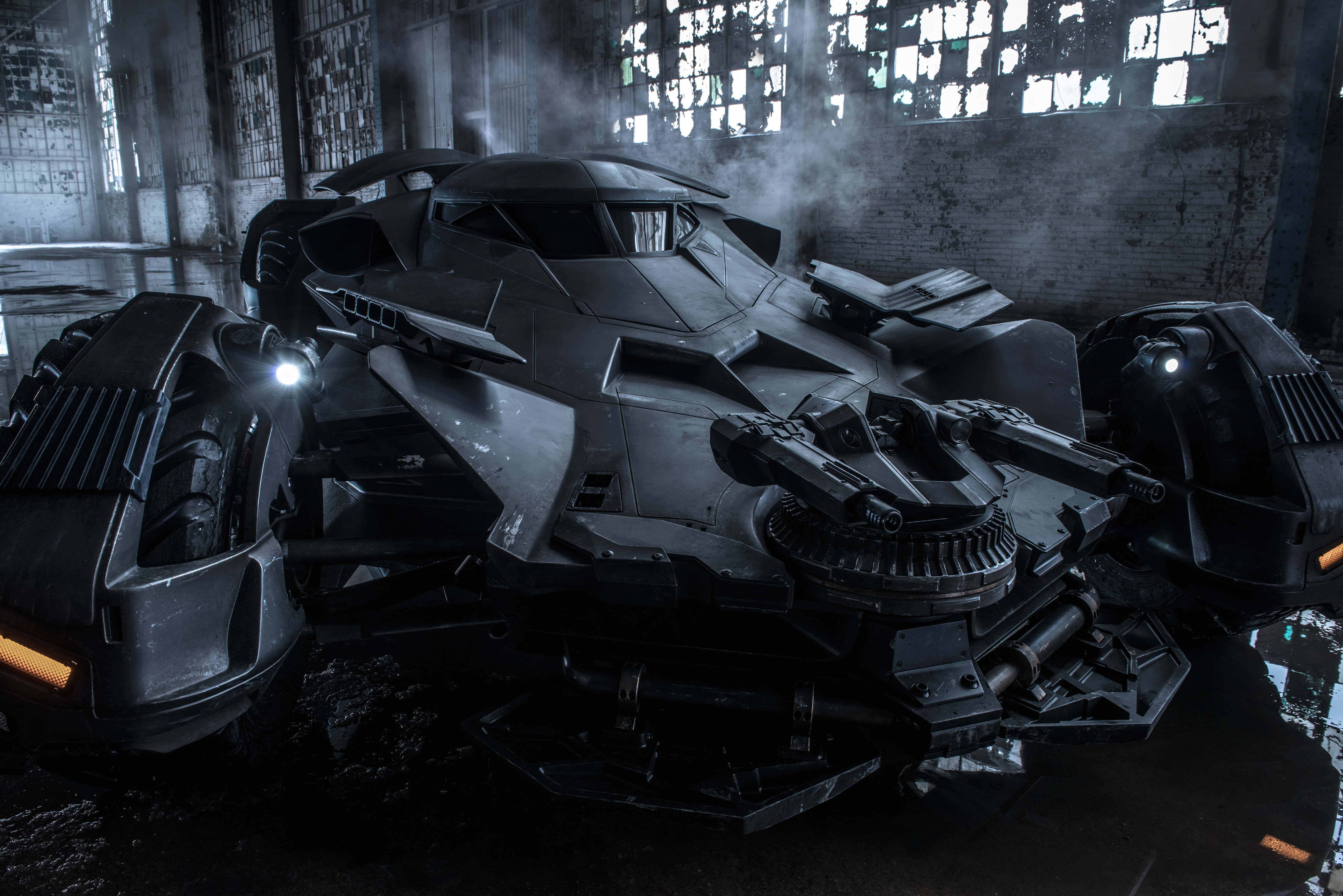 4K Batmobile 桌布 壁紙 — 一架時尚的黑色蝙蝠車停放在煙霧瀰漫的廢棄倉庫中，倉庫有破窗和水窪，映照出其棱角分明的形態。