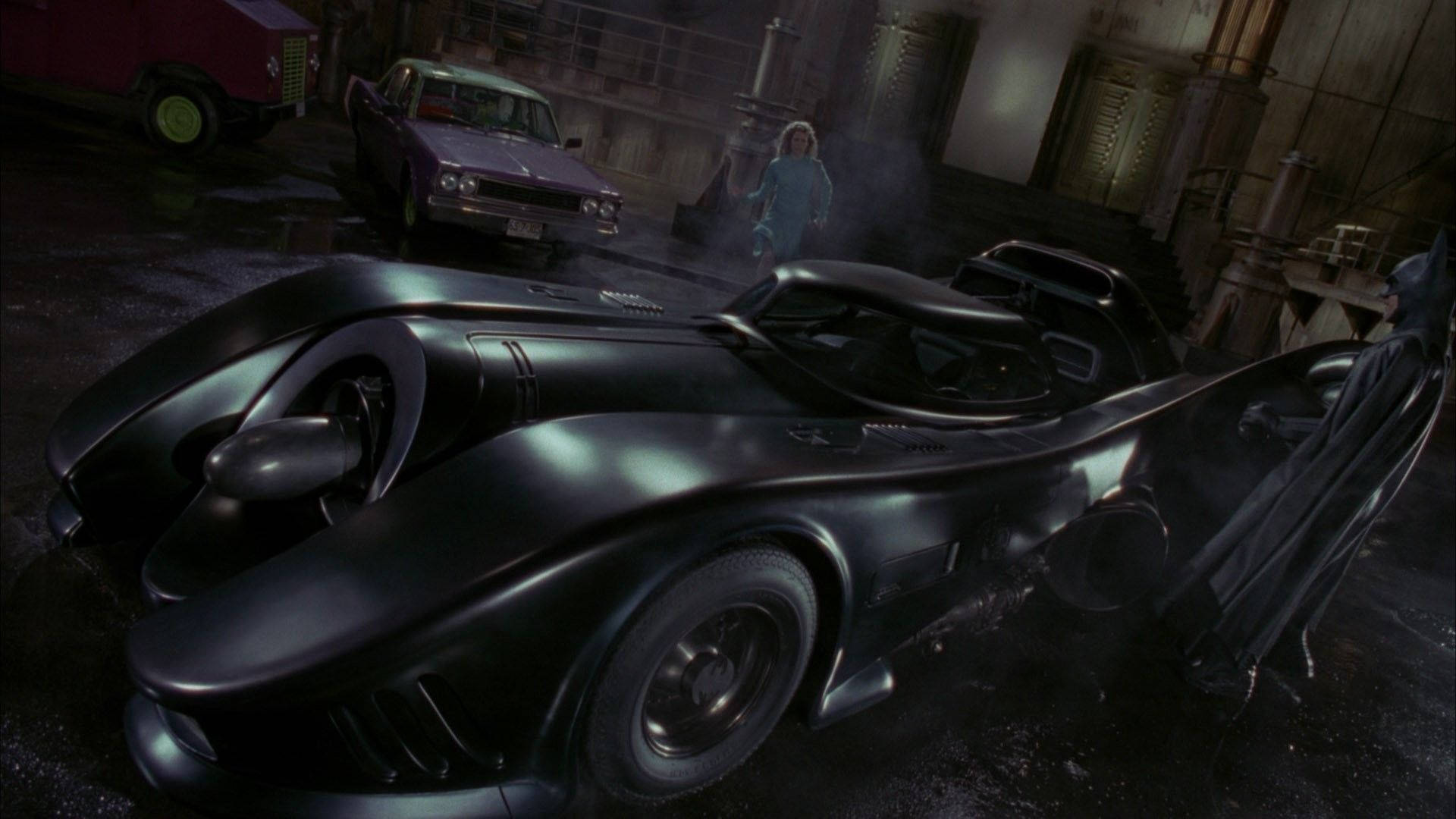 Batmobile 桌布 壁紙 — 一架時尚的黑色蝙蝠車在昏暗燈光下閃耀，位於下雨的工業車庫中，附近站著一位披風人物。