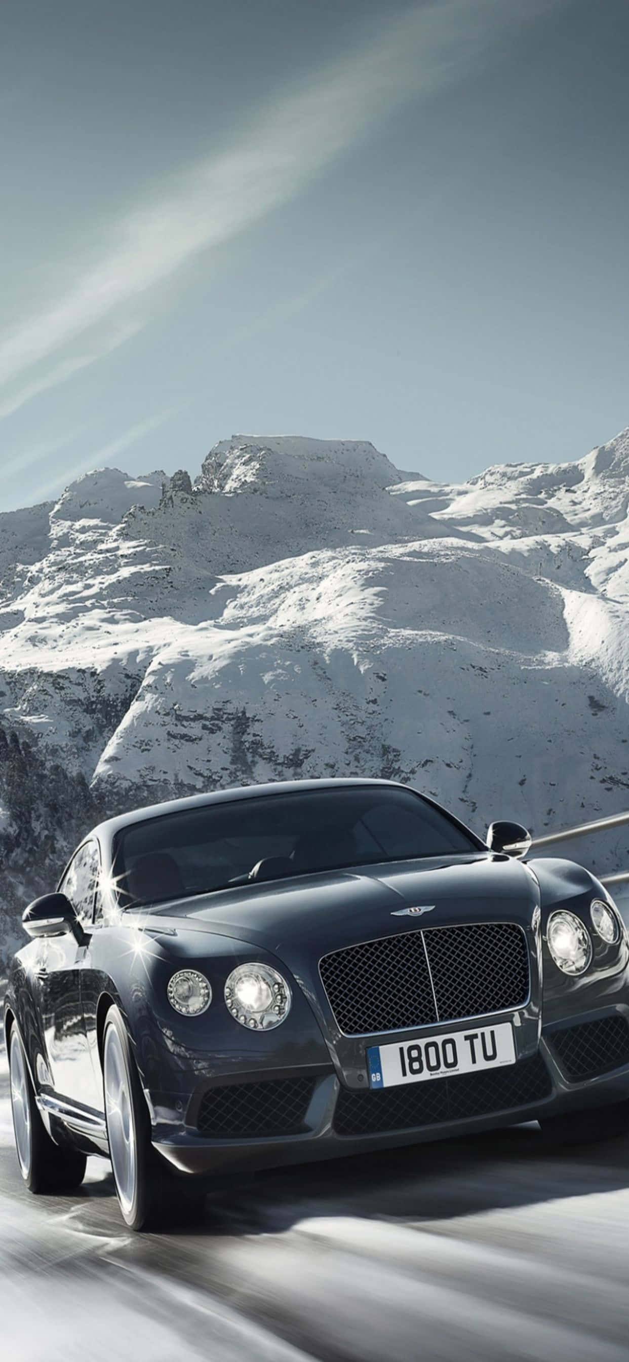 iPhone Xs Max Bentley 桌布 壁紙 — 一架時尚的黑色賓利 Continental GT 在晴朗藍天下，沿著積雪的山路飛馳。