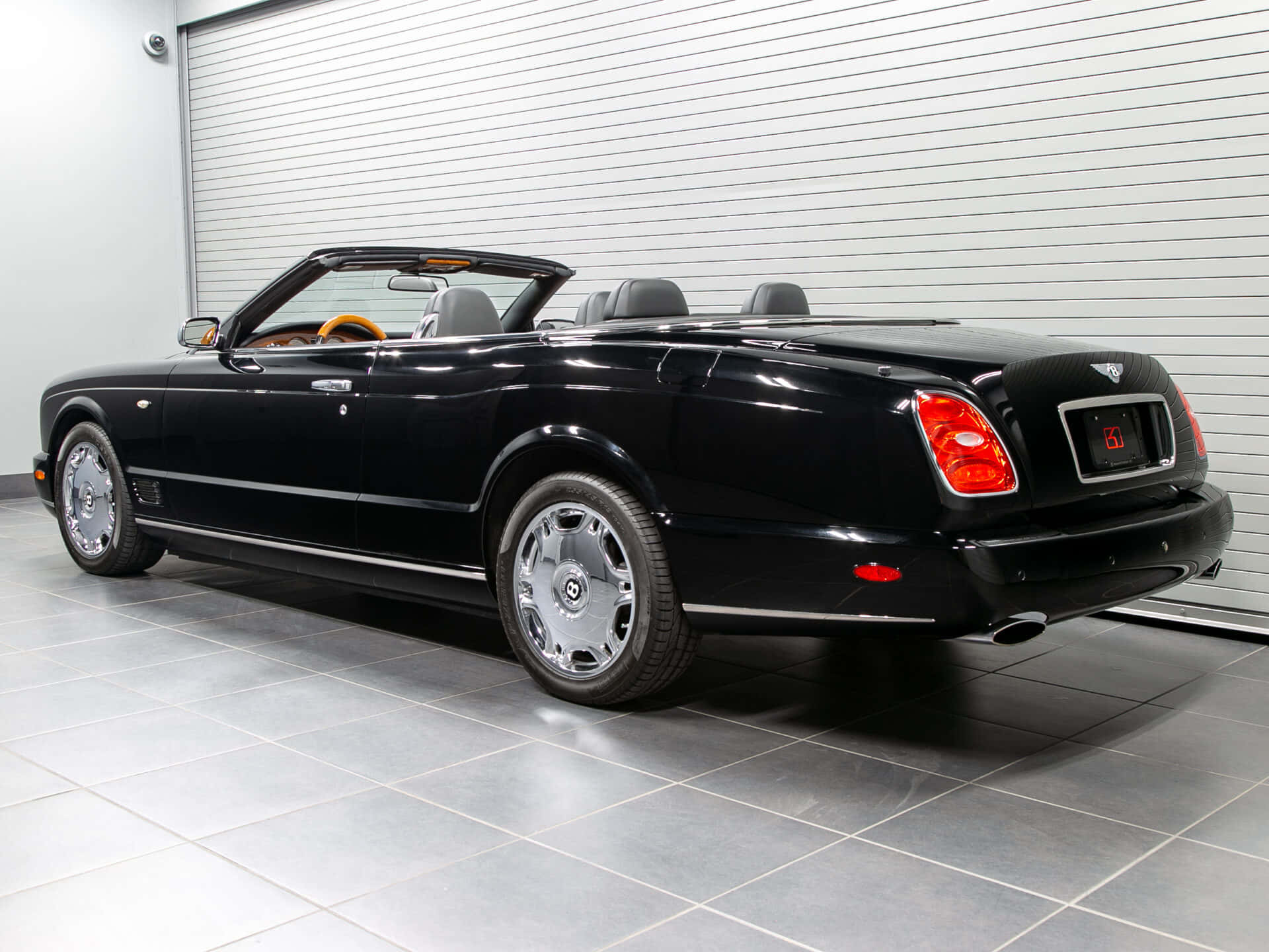 Bentley Azure 桌布 壁紙 — 一架時尚的黑色賓利敞篷車，配備鉻合金輪圈和橙色內裝，停放在一個乾淨的瓷磚車庫中，車庫有白色捲簾門。