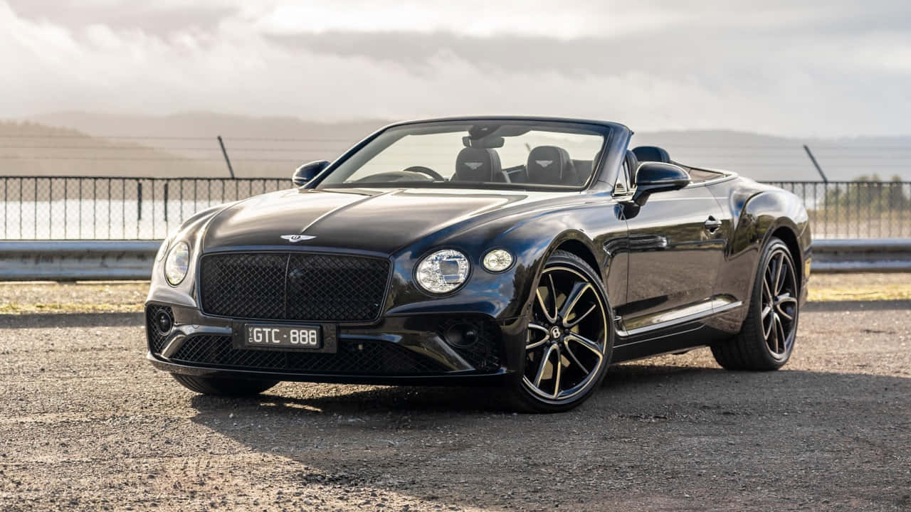 Bentley Continental Gt Convertible 桌布 壁紙 — 一架時尚的黑色賓利敞篷車停泊在碎石路上，旁邊有圍欄觀景點和遠處的山丘，天空多雲。