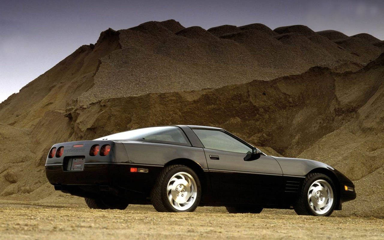 corvette 桌布 壁紙 — 一架時尚的黑色跑車停泊在塵土飛揚的地形上，前方是巨大的岩石山丘，天空柔和。