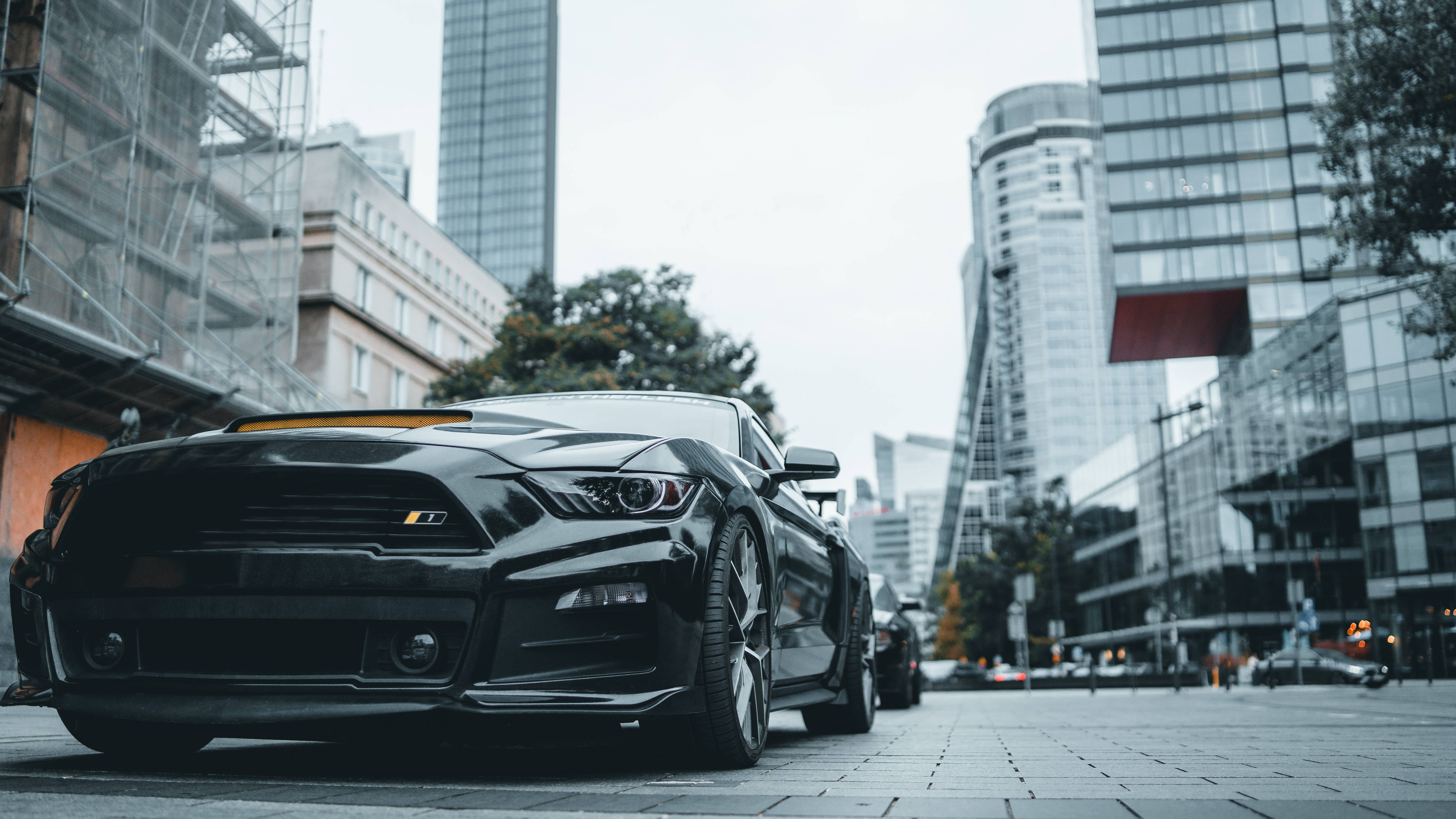 Cool Mustang 桌布 壁紙 — 一架時尚的黑色跑車停泊在現代摩天大樓和建築棚架背景下的城市街道上。