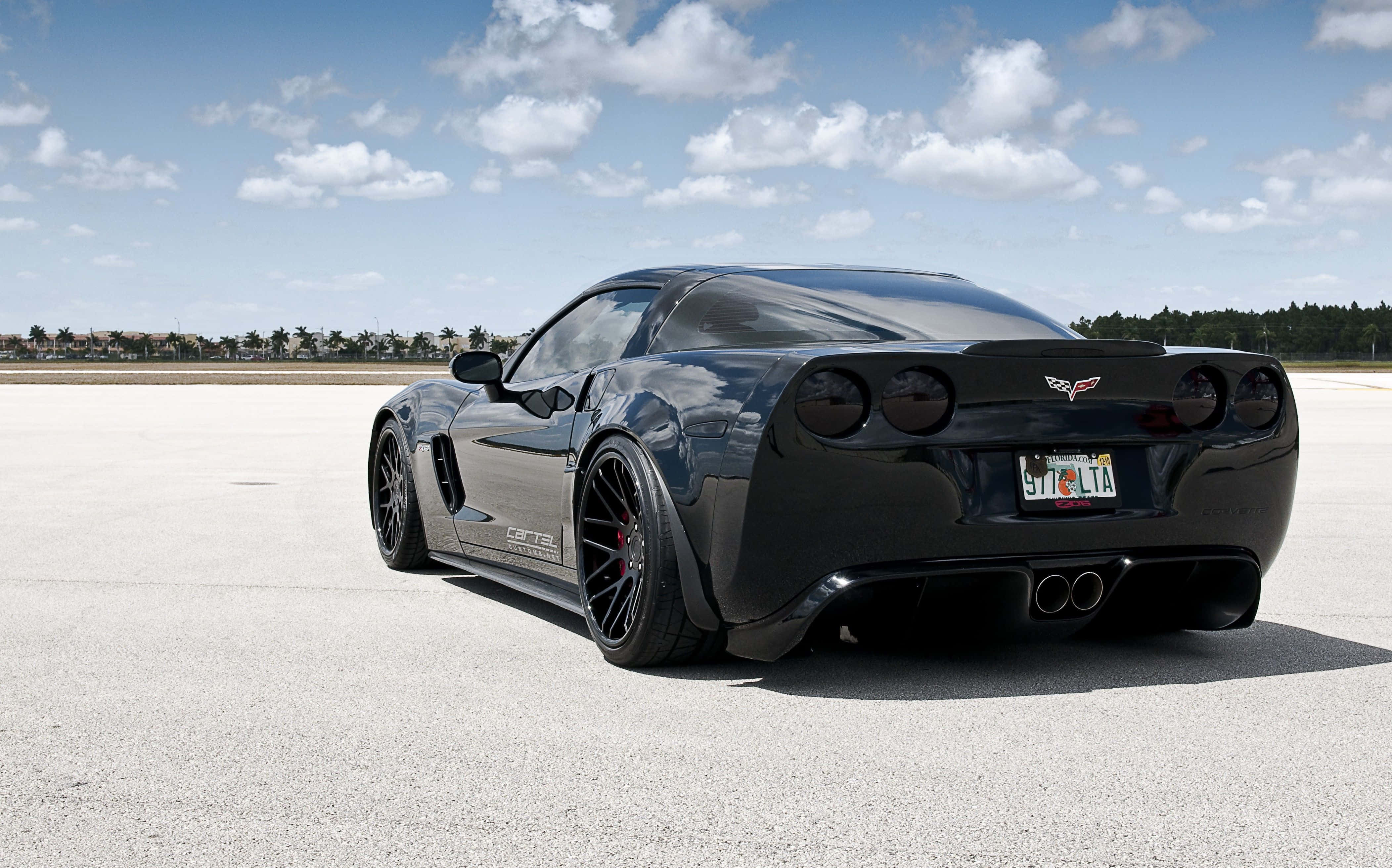 corvette 桌布 壁紙 — 一架時尚黑色Corvette跑車停放在廣闊的混凝土場地上，天空湛藍，白雲點綴。