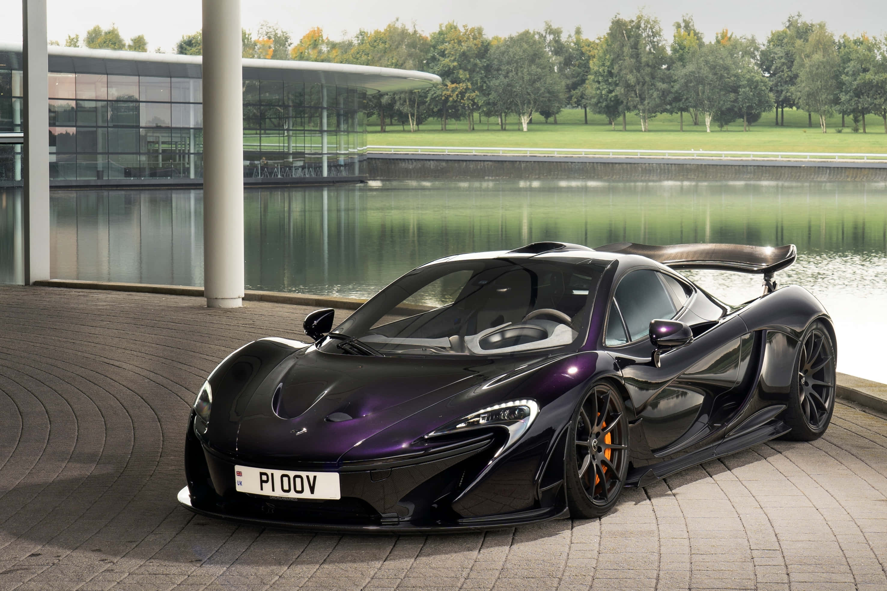 麥拉倫 桌布 壁紙 — 一架時尚黑色McLaren P1跑車帶有紫色亮點，停泊在瓷磚庭院旁，旁邊有現代玻璃建築和寧靜的水面。
