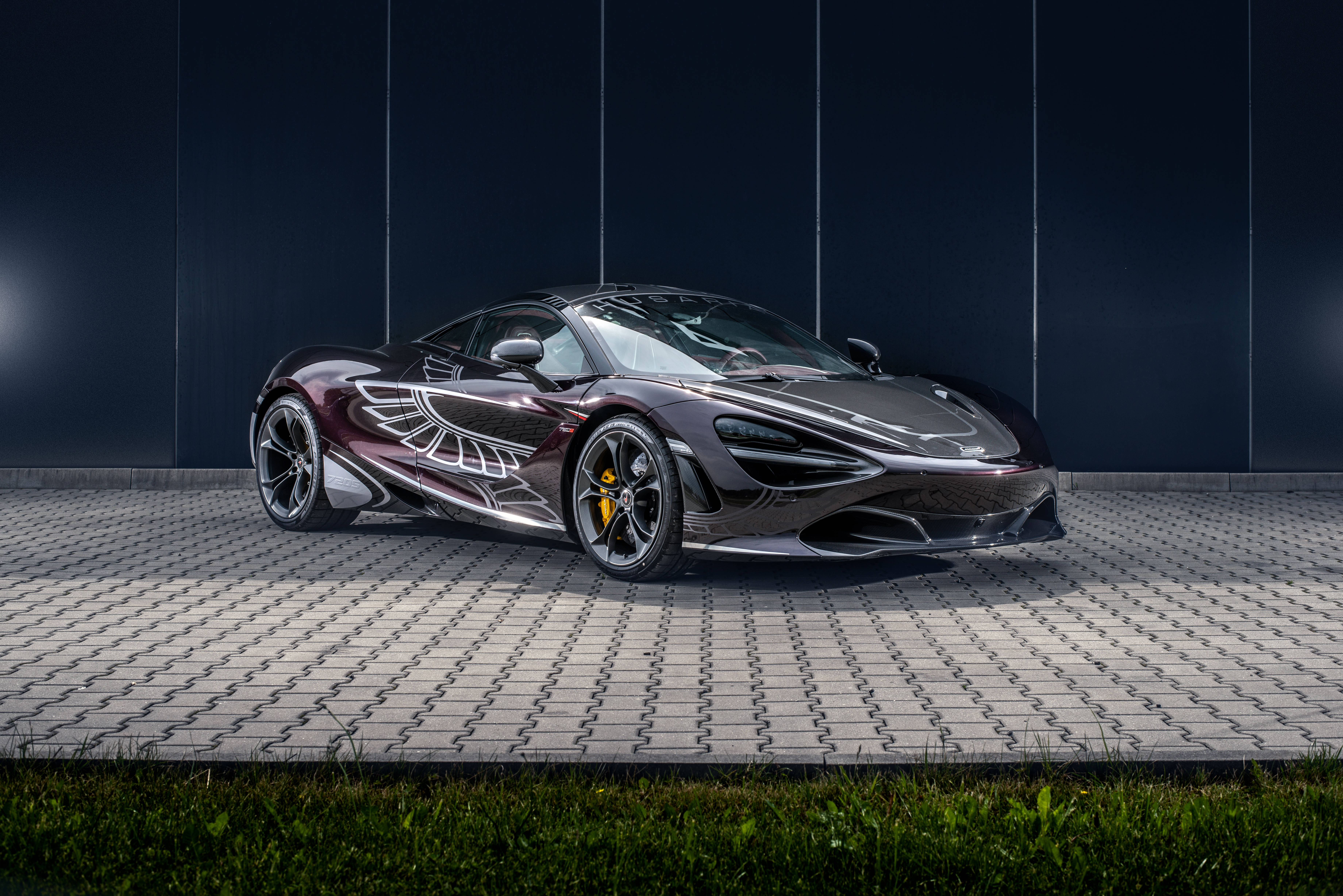 Mclaren 720s 桌布 壁紙 — 一架時尚黑色McLaren跑車帶有銀色翼形貼紙，停放在瓷磚鋪設的路面上，背景是深藍色牆壁。