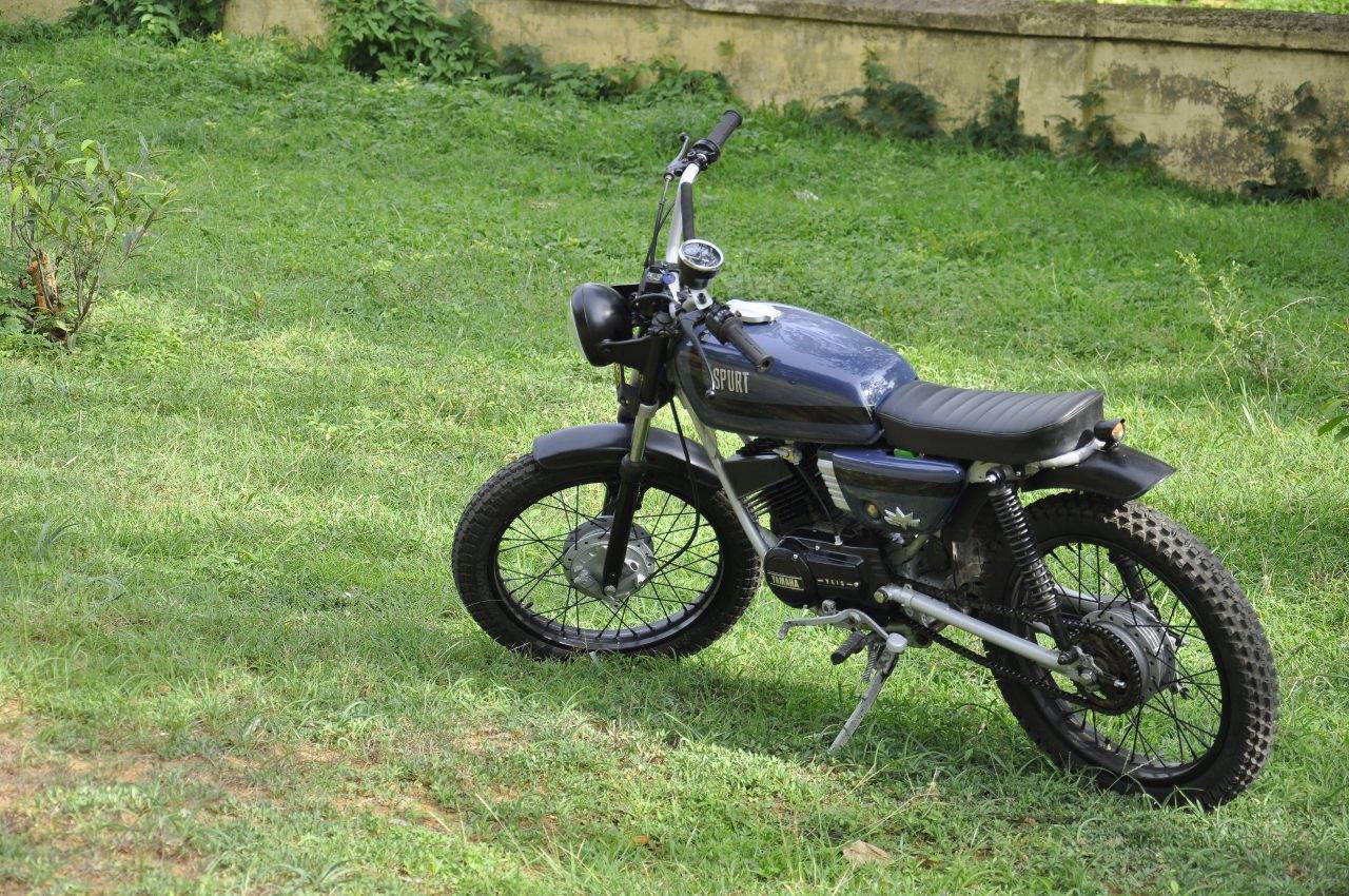 Rx100 桌布 壁紙 — 一架時尚黑色復古機車配備越野輪胎，停放在陽光普照的草地上，旁邊是風化的混凝土牆。