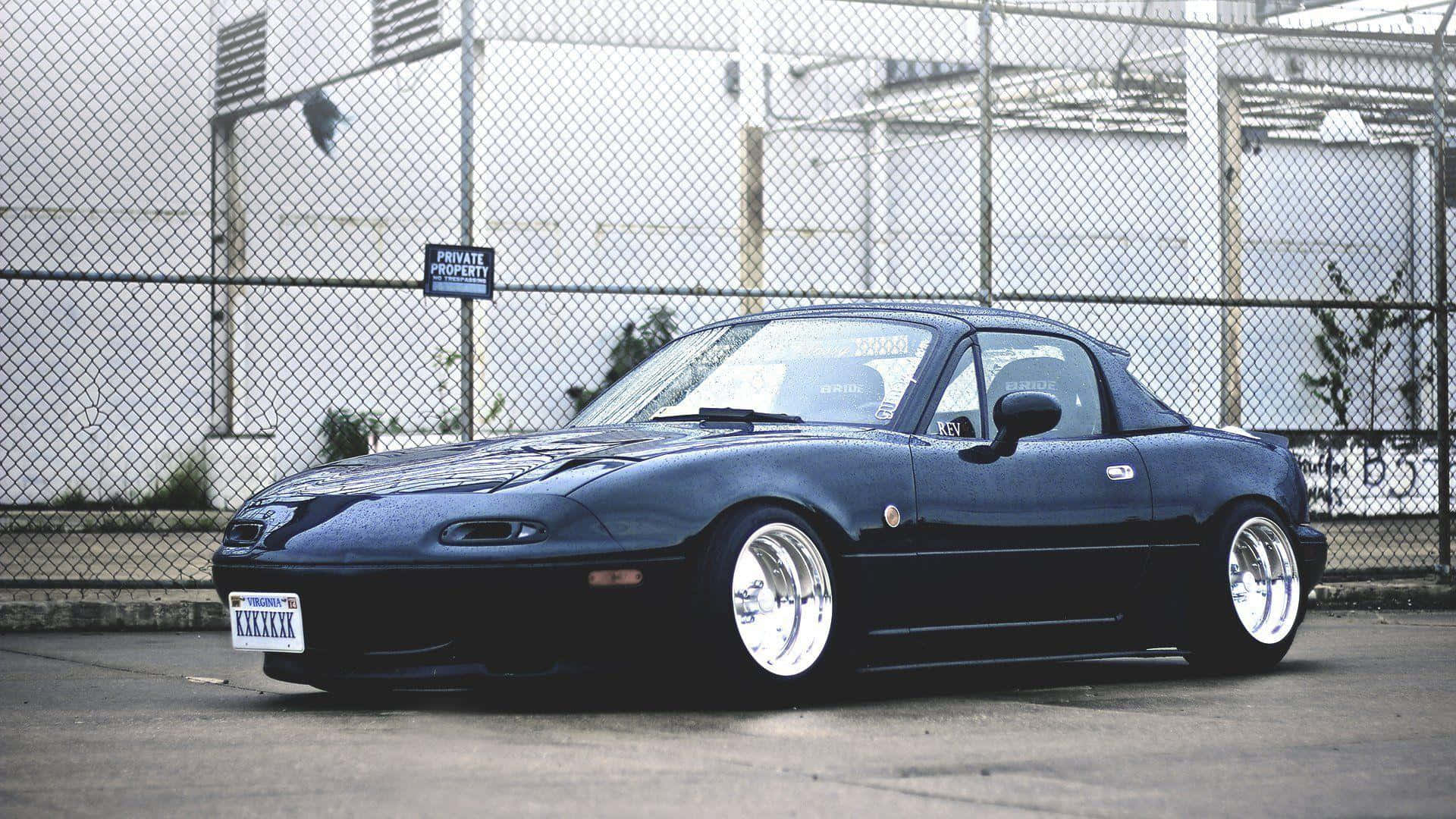 Miata 桌布 壁紙 — 一架時尚黑色改裝Miata跑車配備定制輪圈，在陰天停泊在鐵絲網圍欄後面。
