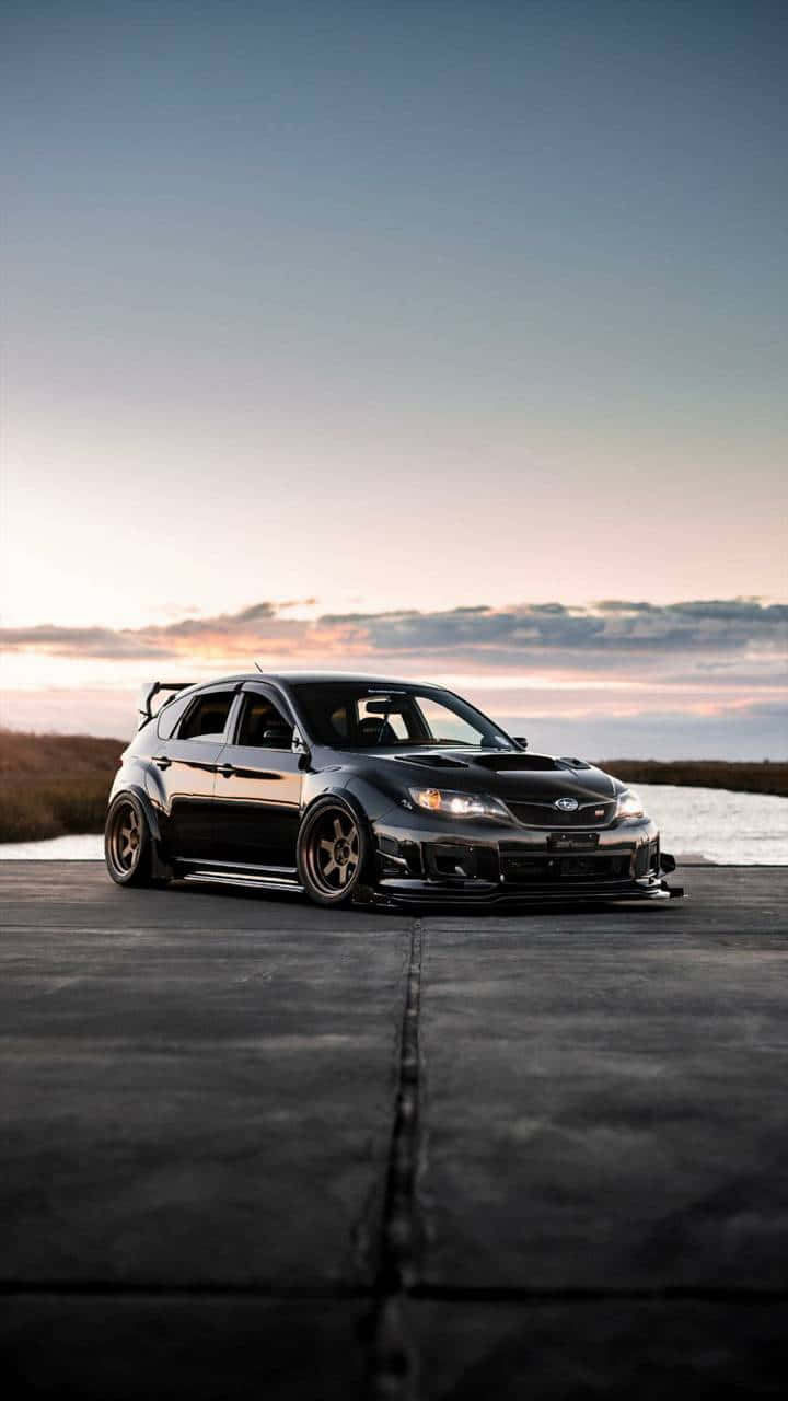 JDM 桌布 壁紙 — 一架時尚黑色改裝Subaru WRX STI在日落時分於混凝土路面上飄移，背景是平靜的水面。