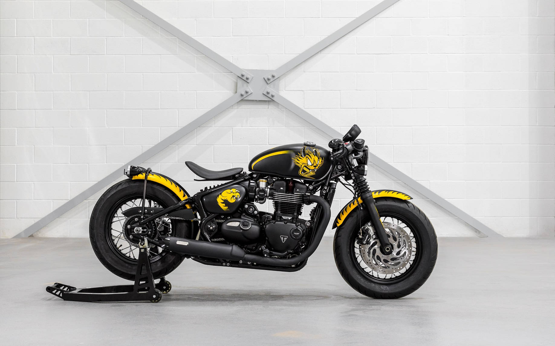 Triumph 桌布 壁紙 — 一架時尚黑色機車帶有搶眼的黃色火焰貼紙，停放在支架上，背景是帶有金屬橫樑的白色磚牆。