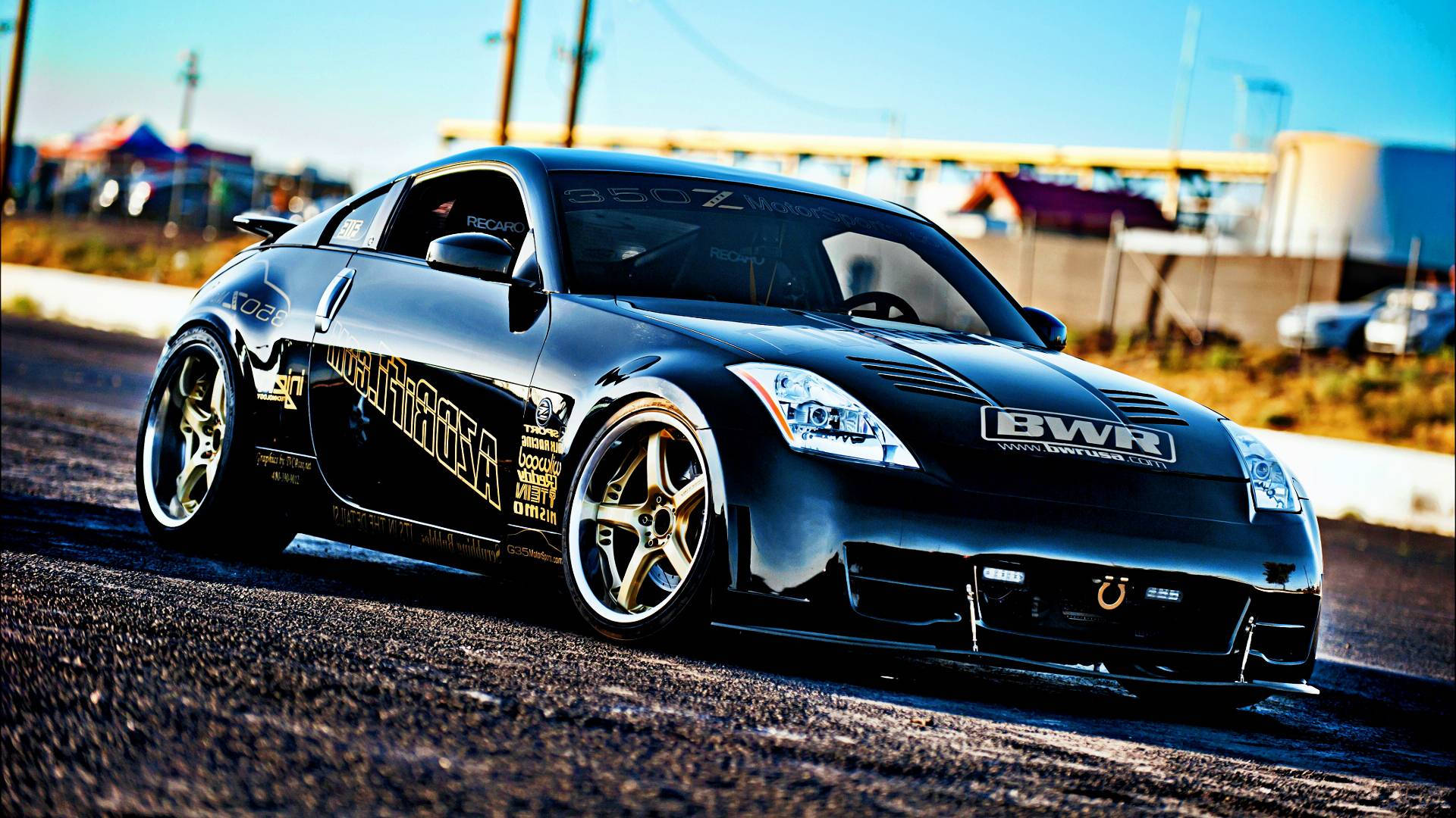 Nissan 350z 桌布 壁紙 — 一架時尚黑色甩尾車，配金色輪圈及贊助商貼紙，在陽光普照的瀝青賽道上奔馳。