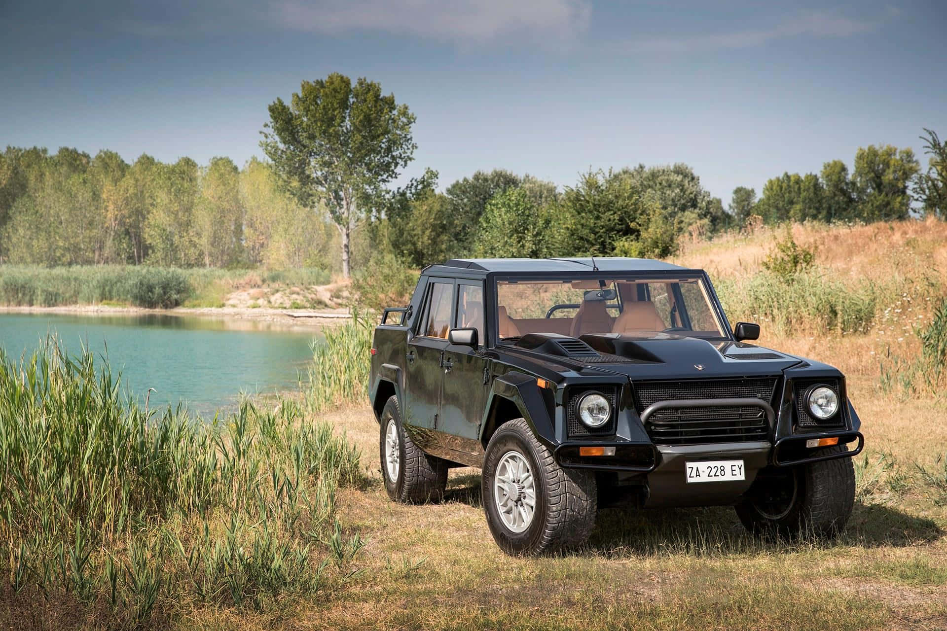 Lamborghini Lm002 桌布 壁紙 — 一架時尚黑色藍寶堅尼LM002 SUV停泊在寧靜的湖邊，四周被高高的草叢和樹木環繞，天空清澈湛藍。
