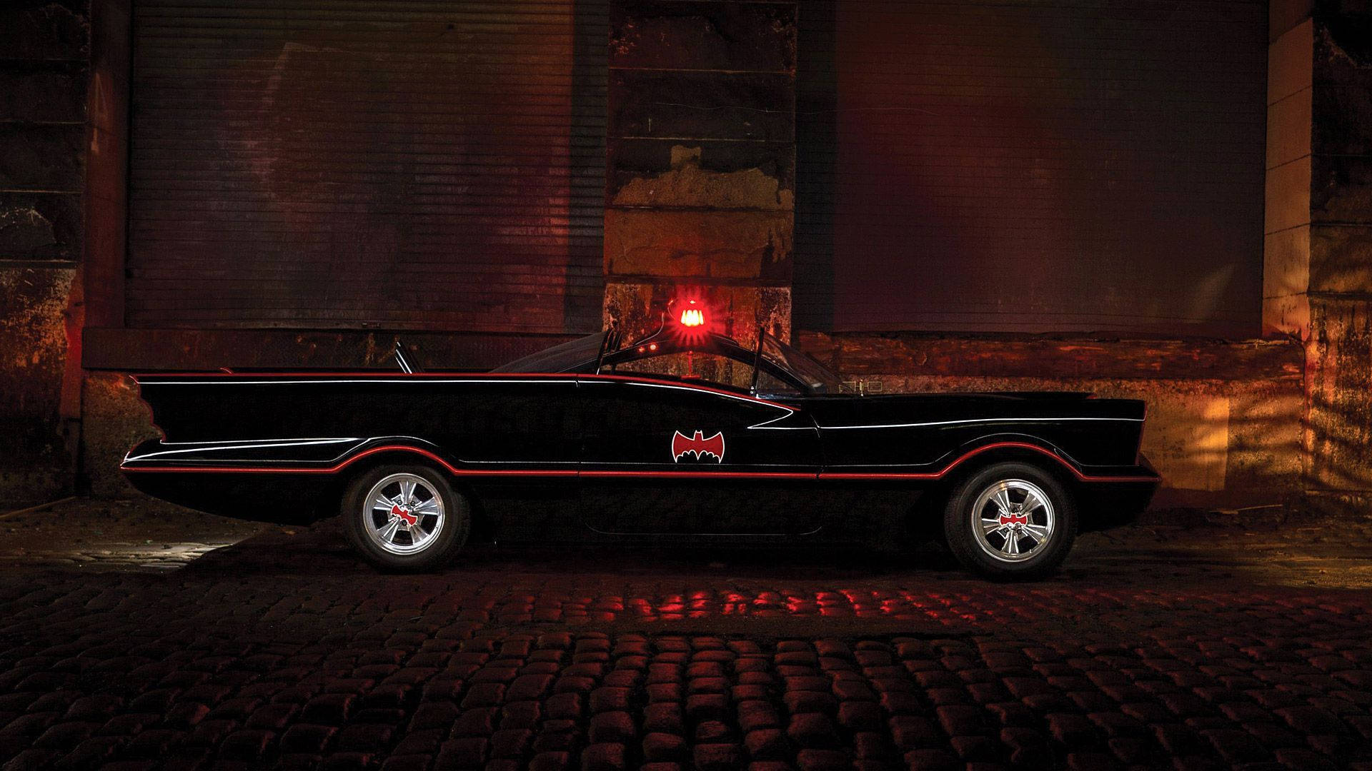 Batmobile 桌布 壁紙 — 一架時尚黑色蝙蝠車，帶有紅色點綴和發光蝙蝠標誌，夜間停在鵝卵石路上，背景是粗獷的城市景觀。