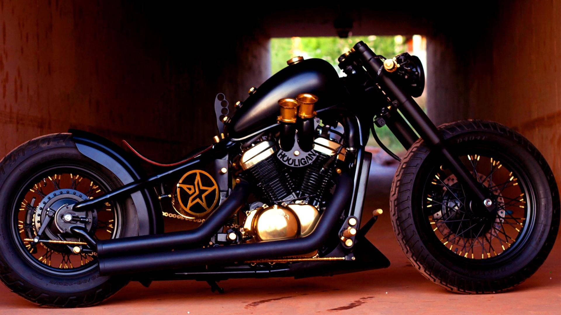 Bobber 桌布 壁紙 — 一架時尚黑色訂製摩托車，帶有金色點綴和星星標誌，停放在隧道下，背景可見綠色植物。