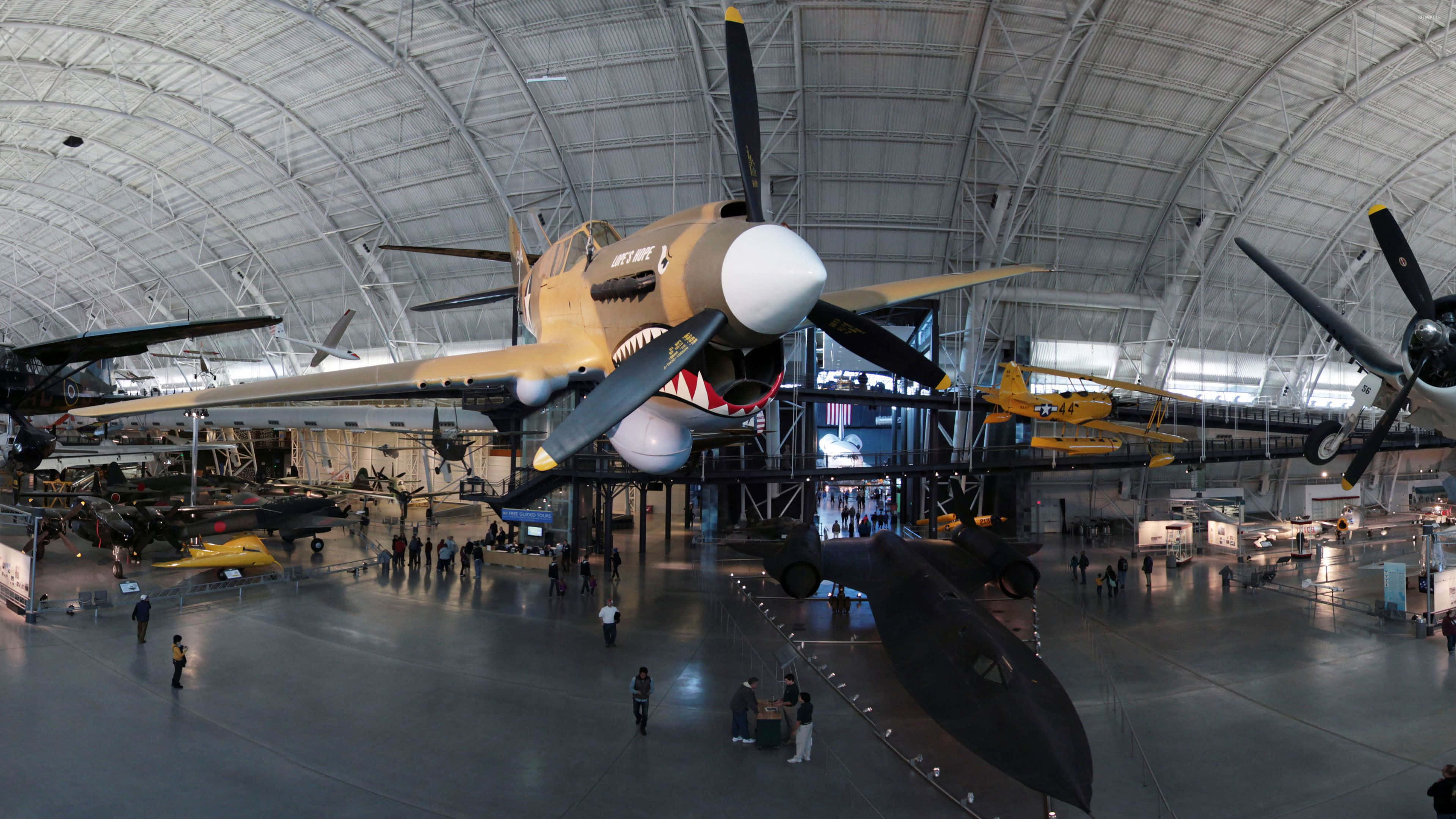 Museums 桌布 壁紙 — 一架機頭繪有鯊魚嘴的復古戰鬥機懸掛在一個寬敞、高天花板的航空博物館中，館內充滿了其他飛機和參觀者。