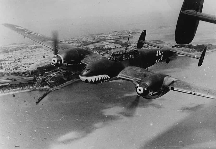 World War 2 桌布 壁紙 — 一架機頭繪有鯊魚嘴的復古軍用飛機，在戲劇性的黑白照片中低空飛過沿海城鎮。