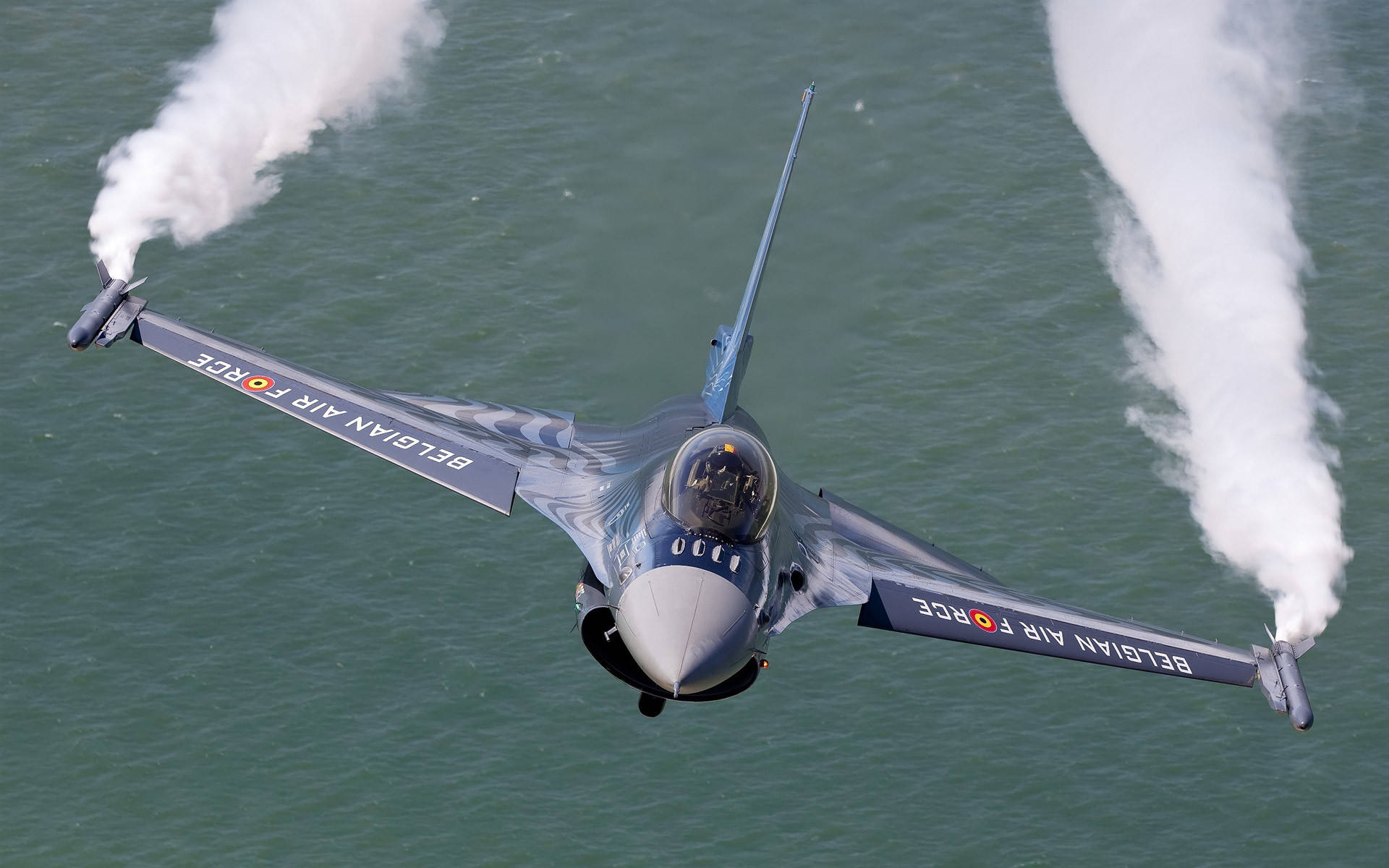 Air Force 桌布 壁紙 — 一架比利時空軍噴射機低飛掠過海洋水面，機翼末端拖出濃厚白色煙霧。