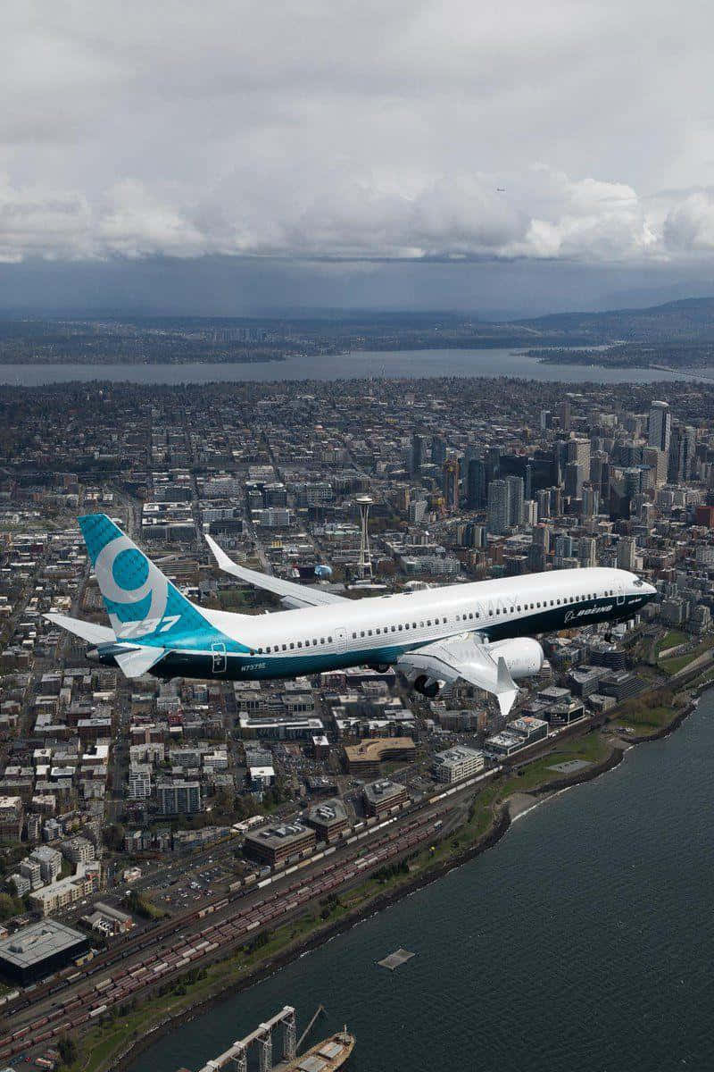 Plane 桌布 壁紙 — 一架波音737飛機，青綠色和白色塗裝，在陰雲密布的天空下翱翔於廣闊的沿海城市景觀之上。